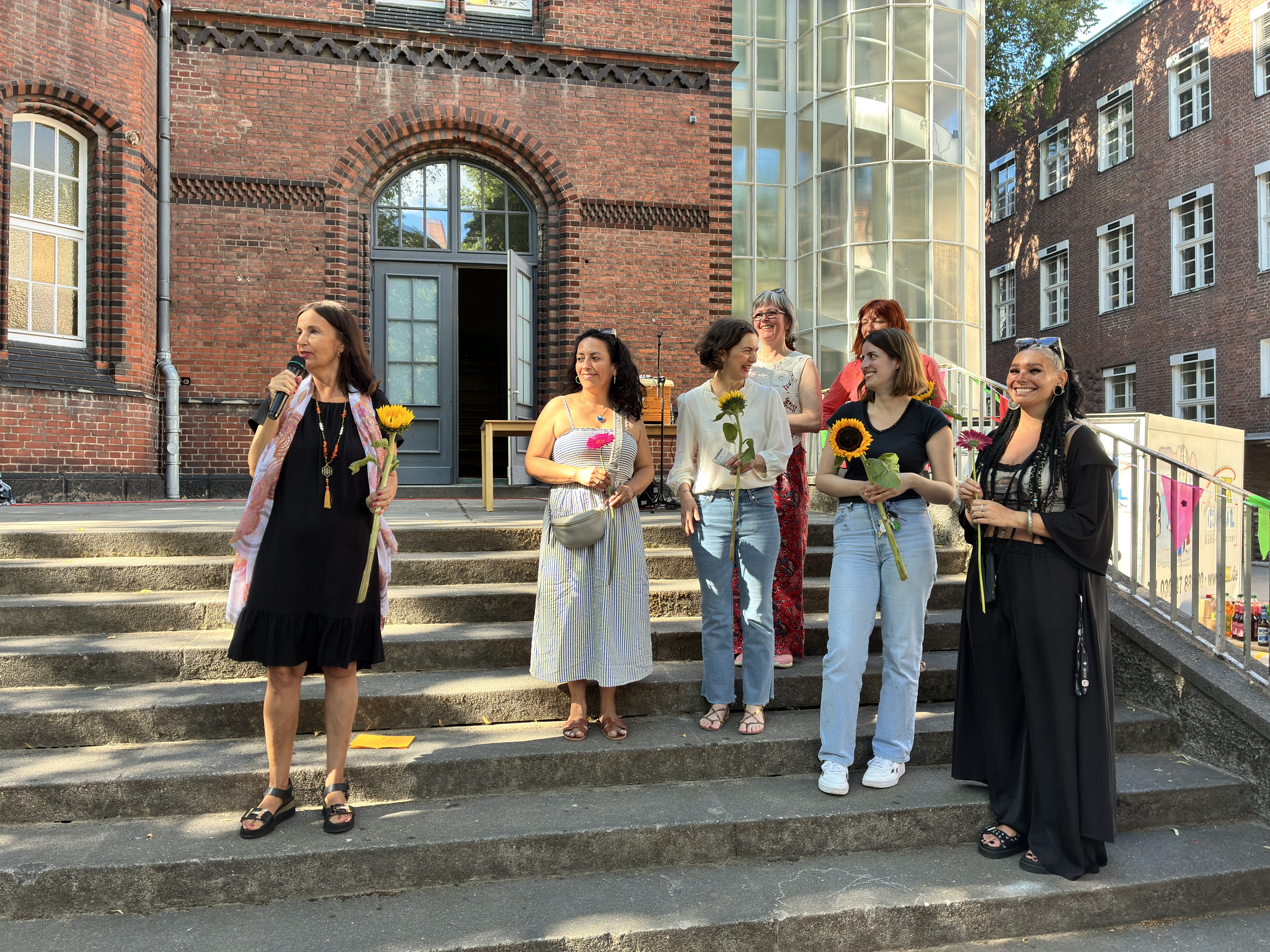 Direktorin Harms hält eine Rede. Neben ihr stehen sechs Frauen, die Sonnenblumen in der Hand halten und lächeln. Im Hintergrund sieht man ein Backsteingebäude.