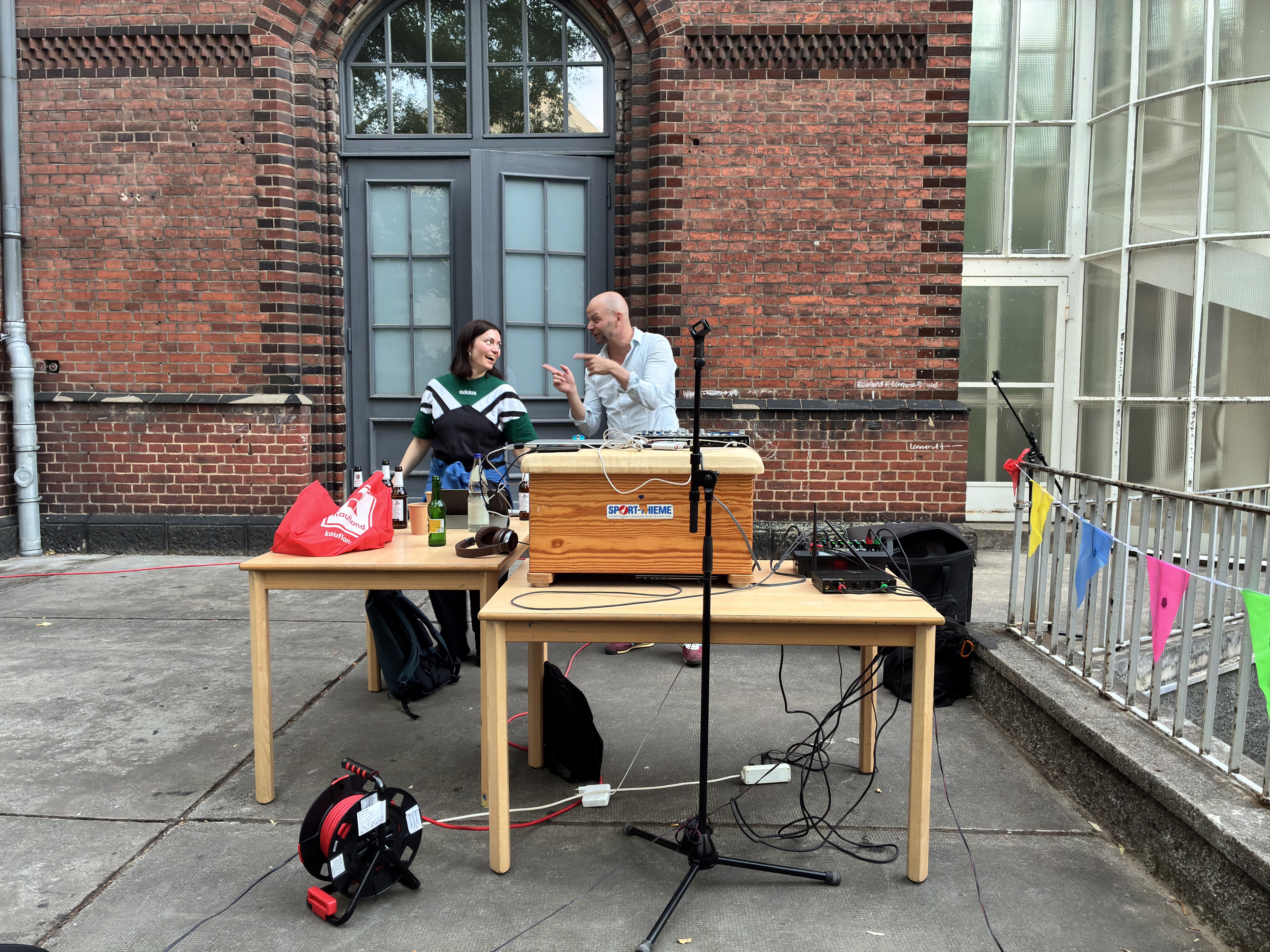 Eine Frau und ein Mann stehen hinter einem Tisch, auf dem ein DJ-Pult aufgebaut ist und lachen. Im Hintergrund sieht man ein Backsteingebäude und an der Seite eine bunte Wimpelkette. 