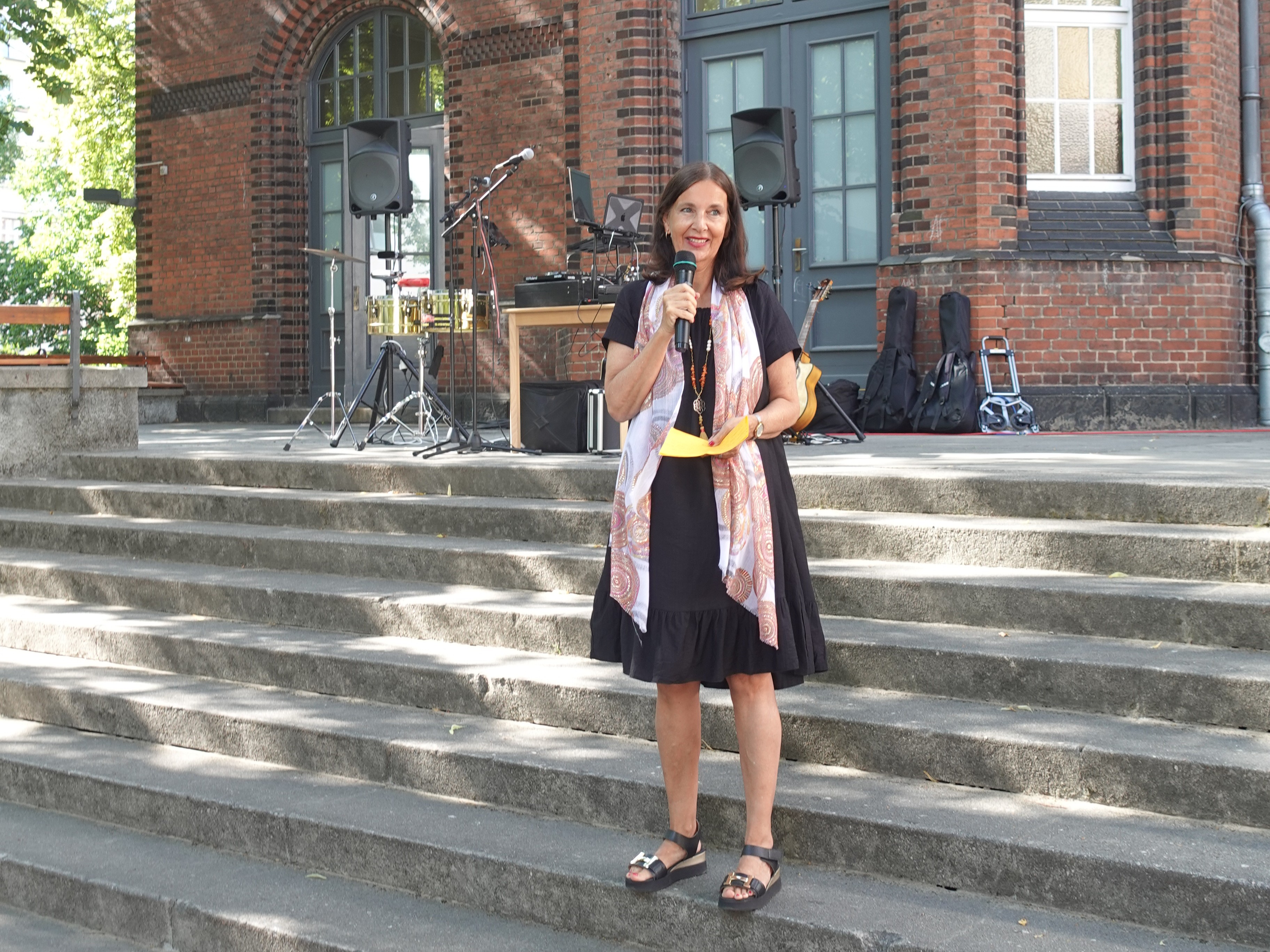 Henriette Harms, Direktorin des Pestalozzi-Fröbel-Hauses, steht auf einer Treppe und hält eine Rede. Sie hat ein Mikrofon in der Hand und lächelt. Im Hintergrund sieht man ein Backsteingebäude. 