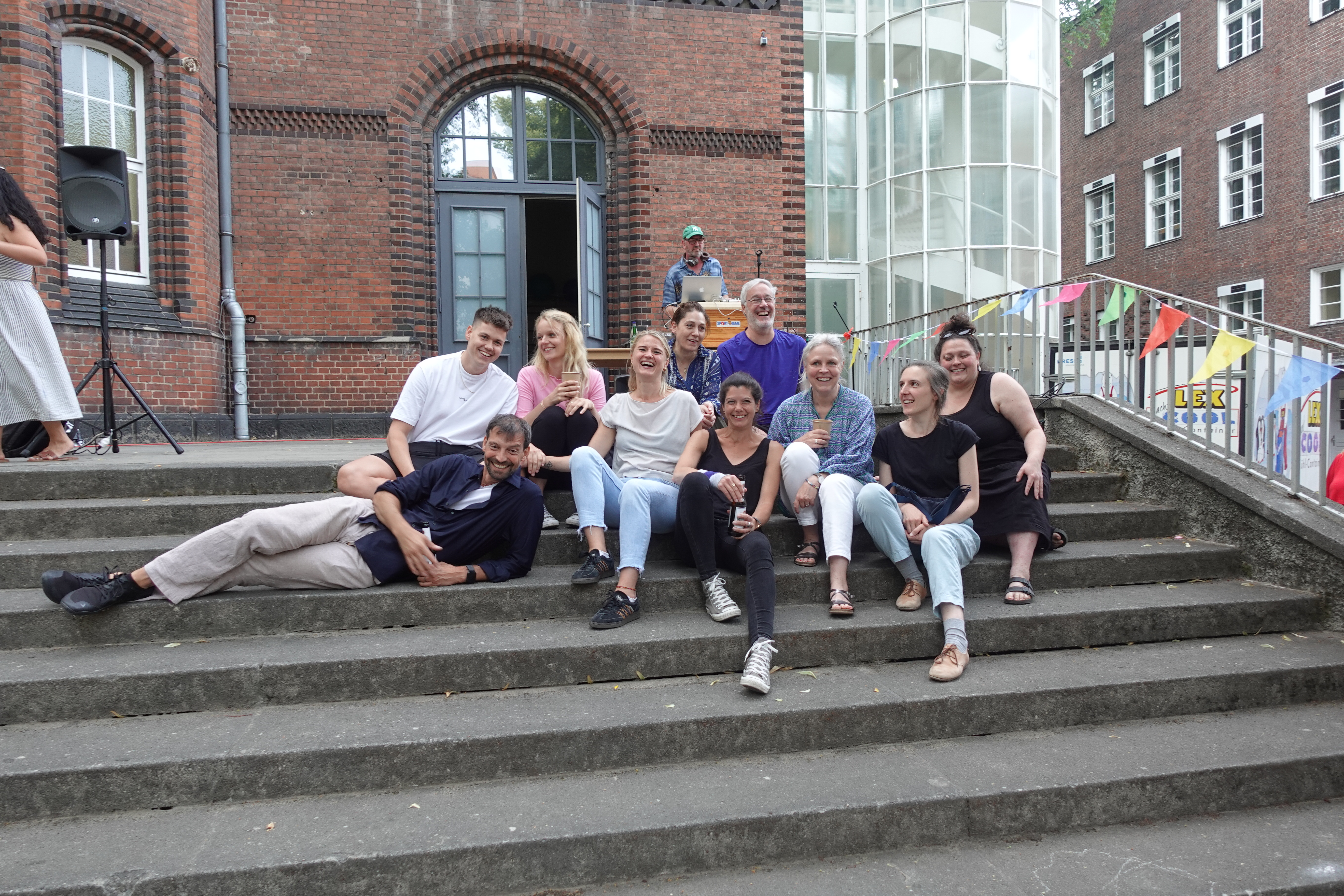 Elf erwachsene Menschen sitzen auf einer Treppe und lachen. Im Hintergrund sieht man einen Mann am DJ Pult, der Musik auflegt. 