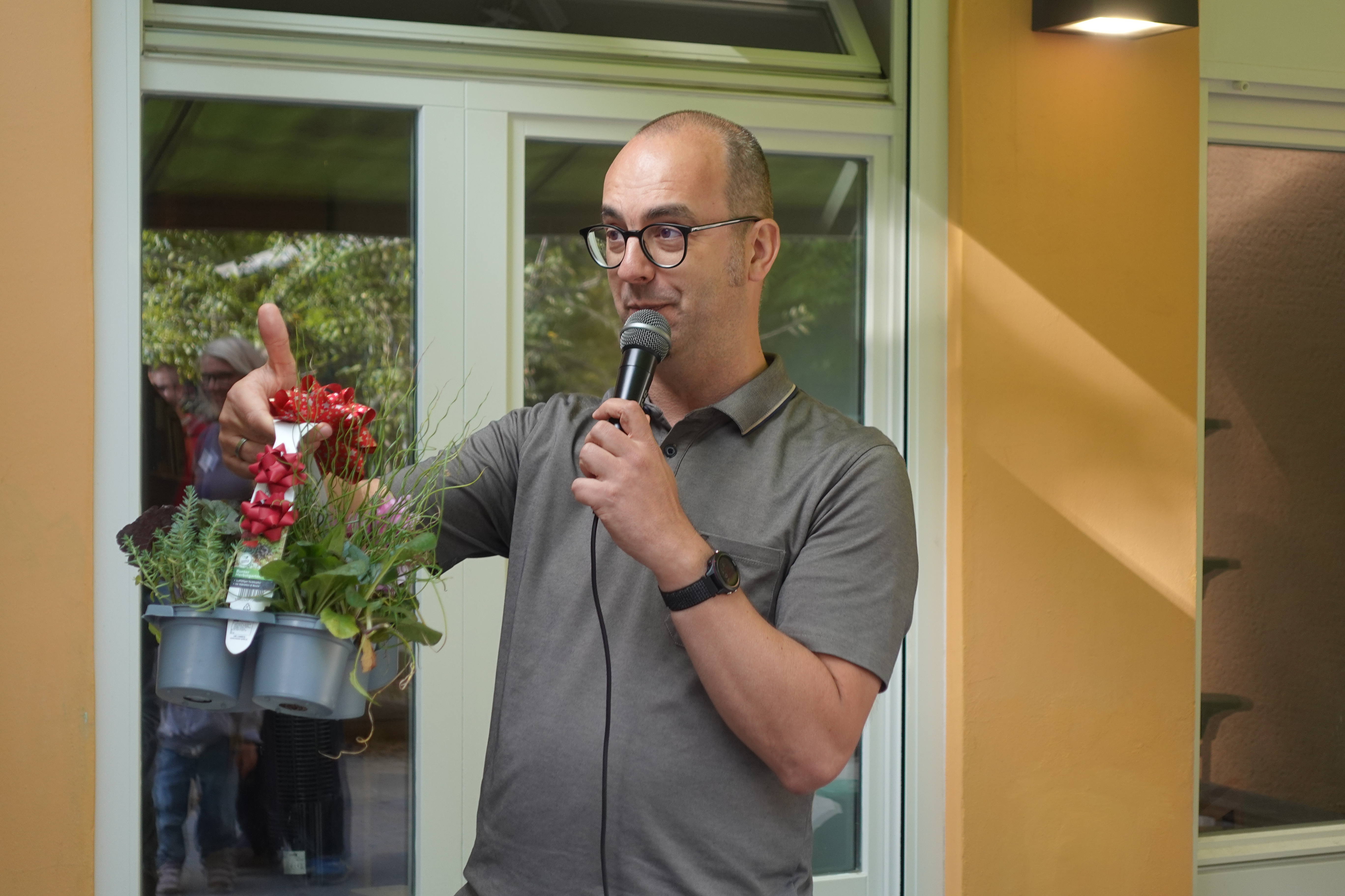 Ein Mann mit Brille spricht in ein Mikrofon und hält einen Topf mit Pflanzen in der anderen Hand. Er steht vor einer gelben Wand mit Fenster im Hintergrund.