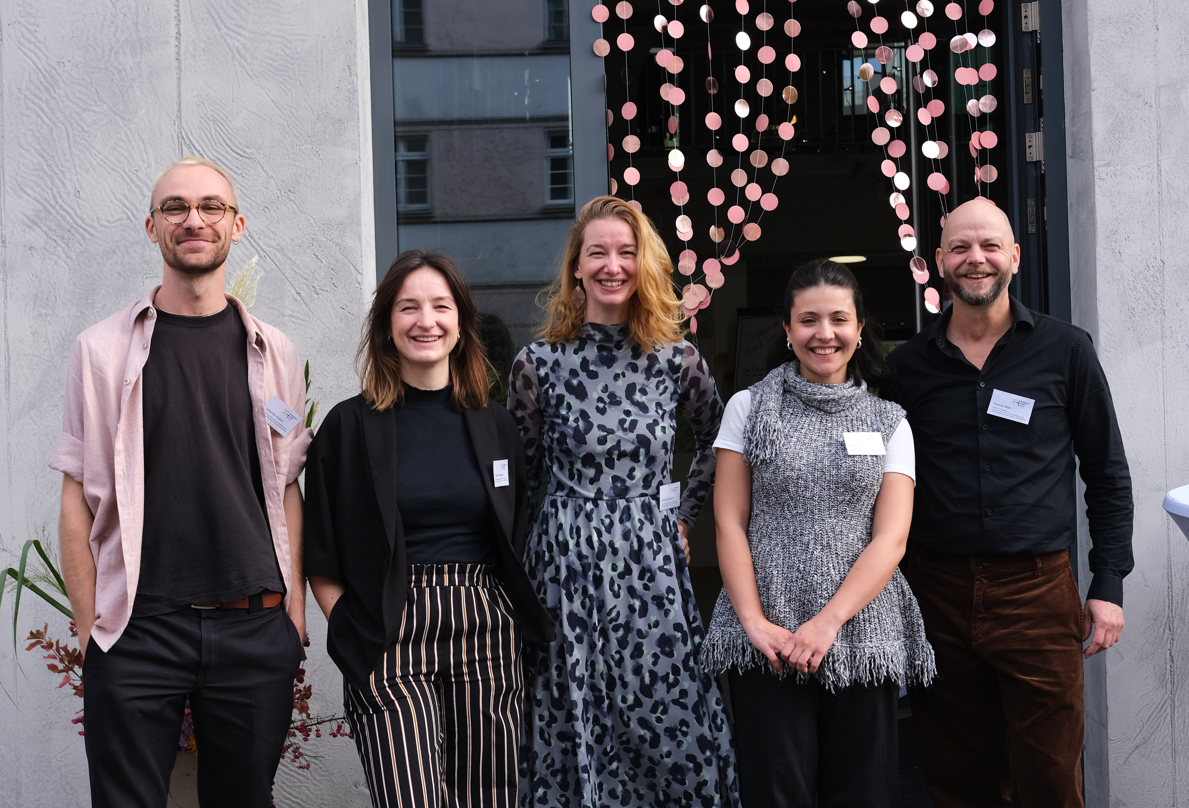 Fünf Personen posieren für ein Foto vor einem Eingang mit dekorativen Lichtern. Sie tragen verschiedene Outfits, die von casual bis elegant reichen.