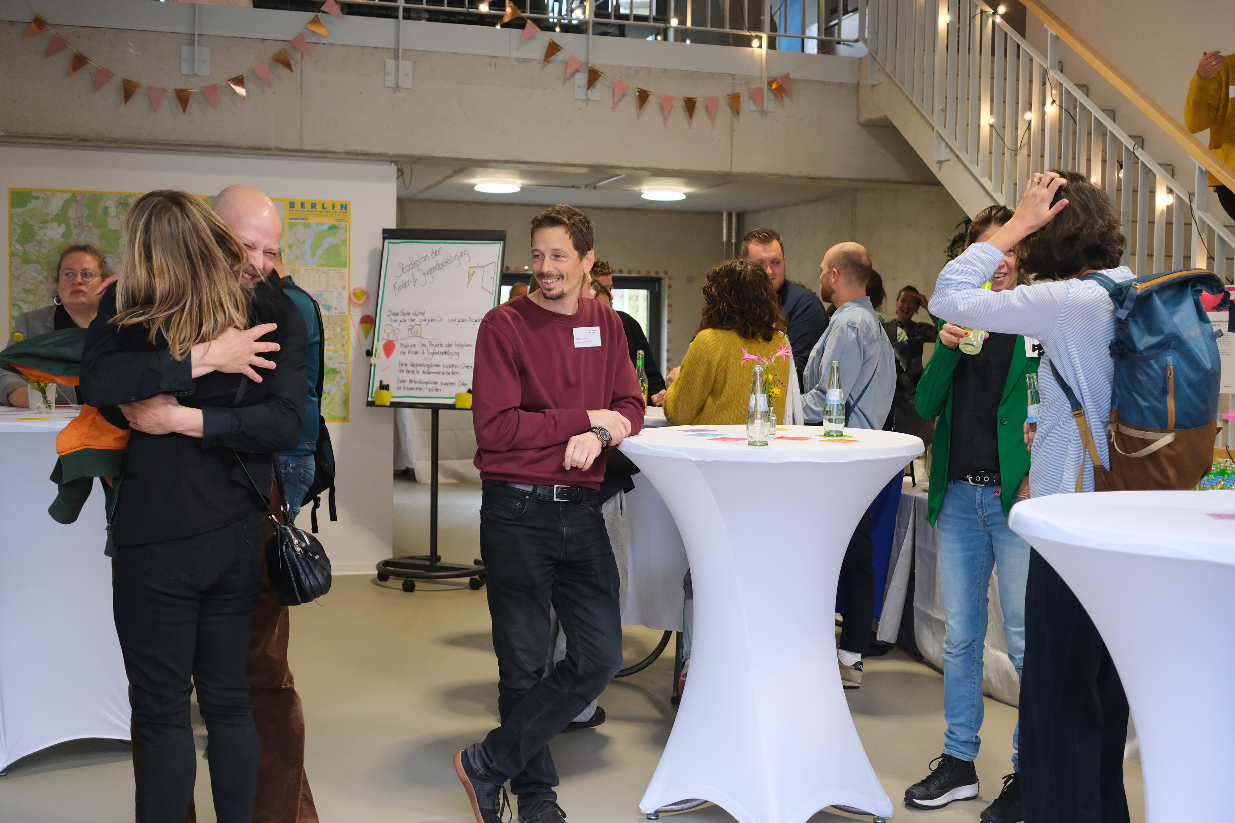 Mehrere Personen stehen um Stehtische in einem Raum mit bunten Dekorationen. Einige Personen umarmen sich, während andere Getränke in der Hand halten. An einer Wand sind Plakate und ein Flipchart sichtbar.