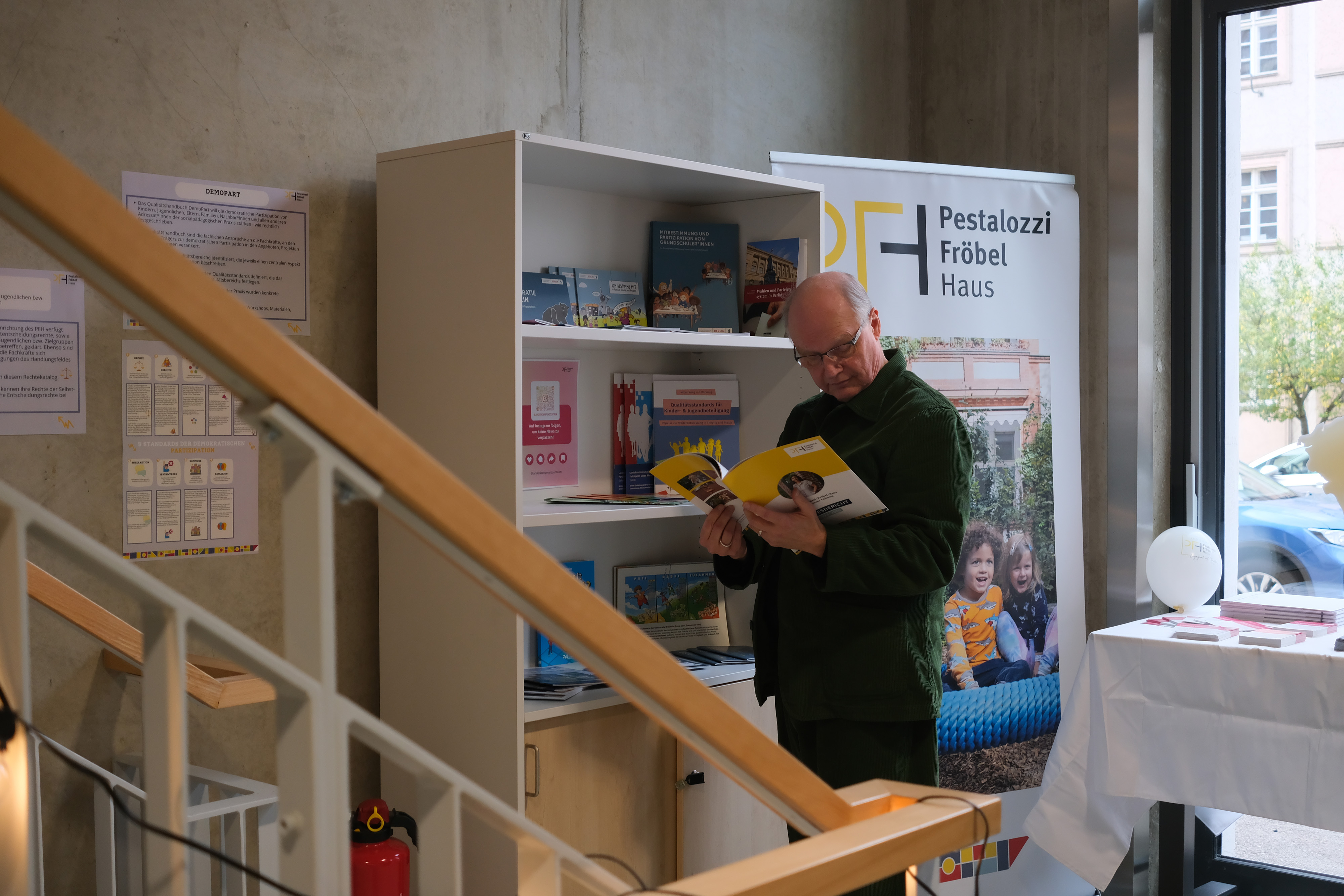 Ein Mann steht an einem Regal in einem modernen Raum und betrachtet Broschüren. Im Hintergrund sind weitere Informationsmaterialien und ein Banner mit dem Logo "Pestalozzi Fröbel Haus" sichtbar.