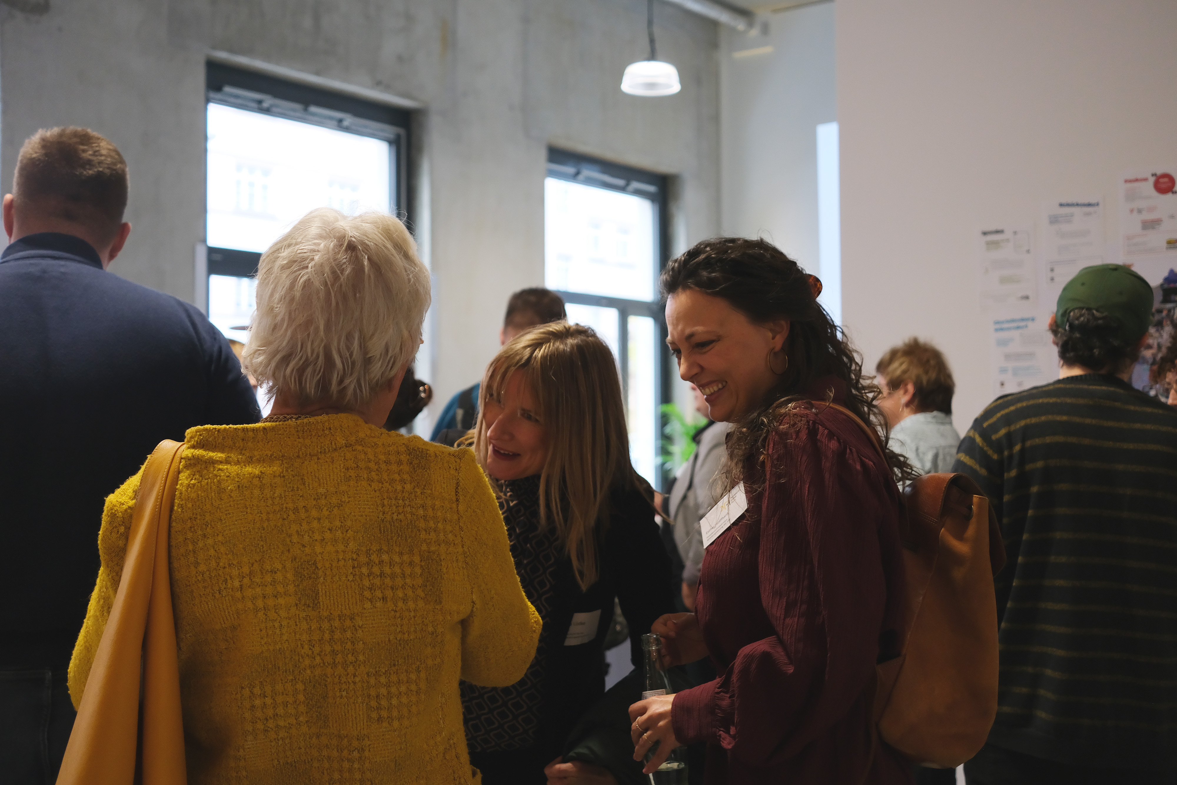 Eine Gruppe von Personen steht in einem Raum, während sie miteinander sprechen. Eine Frau mit kurzen, blonden Haaren trägt einen gelben Pullover und hat eine Tasche über die Schulter. Im Hintergrund sind weitere Personen und Fenster sichtbar.