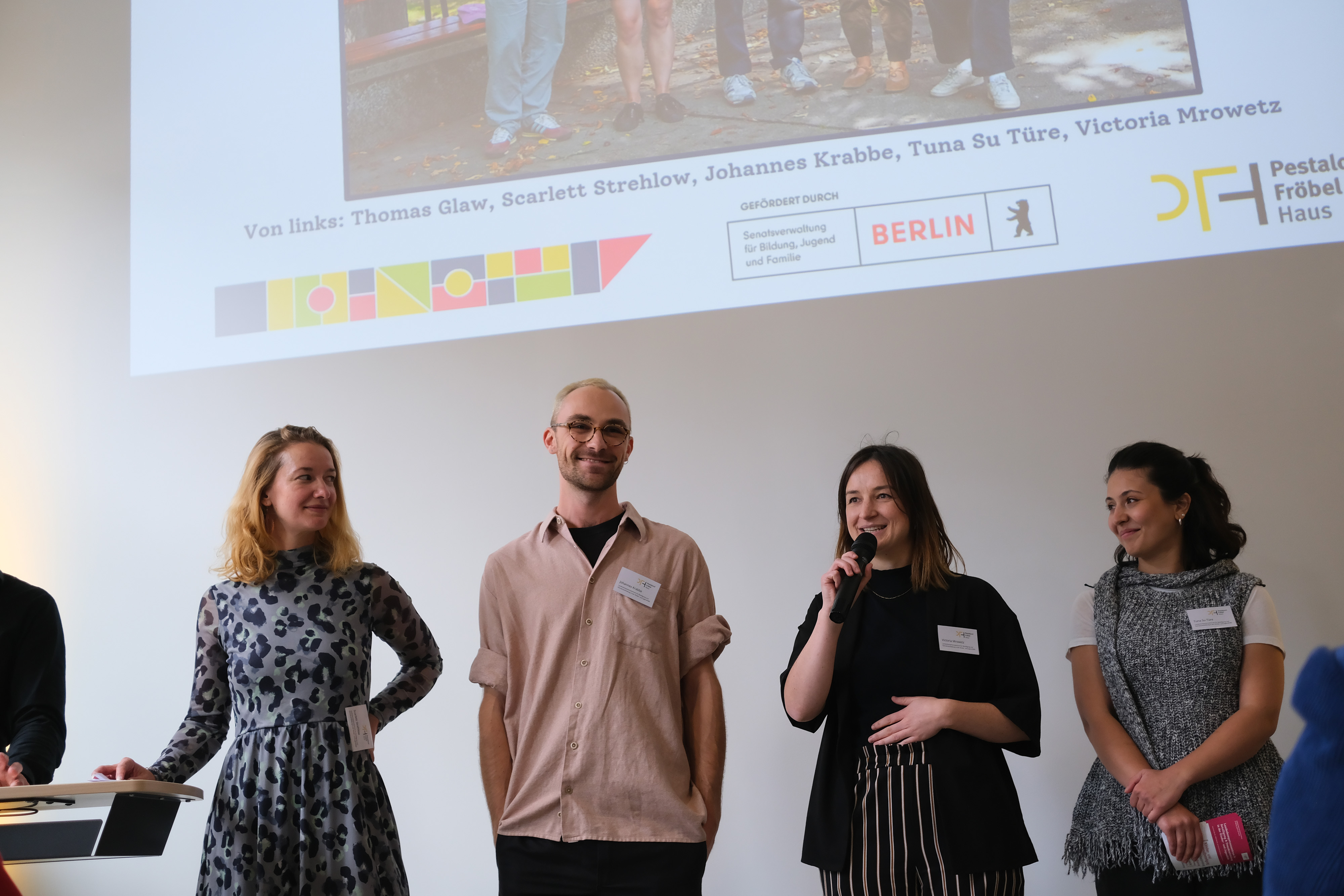 Eine Gruppe von fünf Personen steht auf einer Bühne vor einem großen Bildschirm, auf dem eine Präsentation angezeigt wird. Die Personen halten Mikrofone und scheinen bereit zu sein, etwas zu präsentieren.