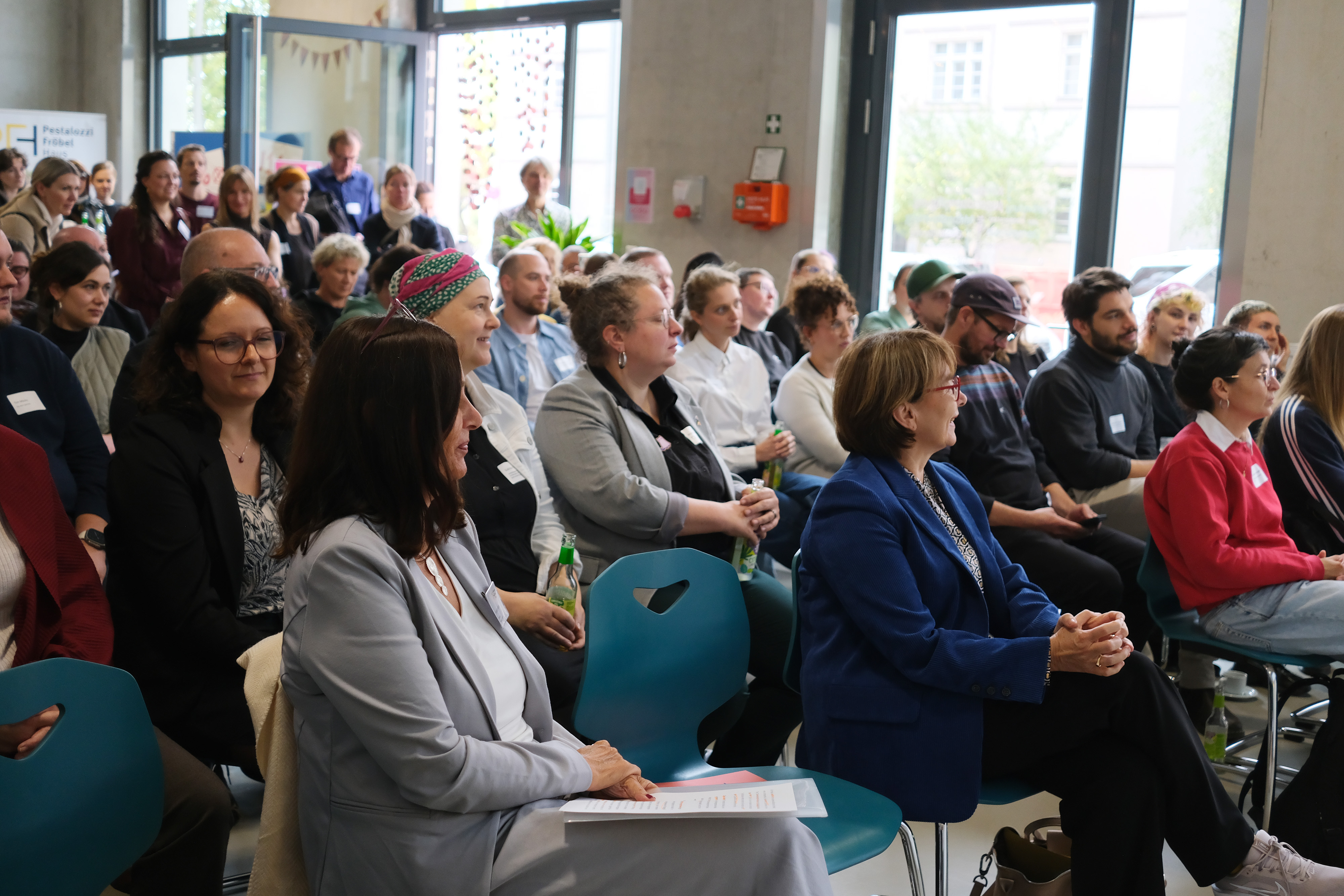 Eine Gruppe von Menschen sitzt in einem Raum mit großen Fenstern und einer modernen Einrichtung. Einige Personen tragen Namensschilder. Im Vordergrund sind zwei Frauen zu sehen, eine in einem grauen Blazer und die andere in einem blauen Anzug.