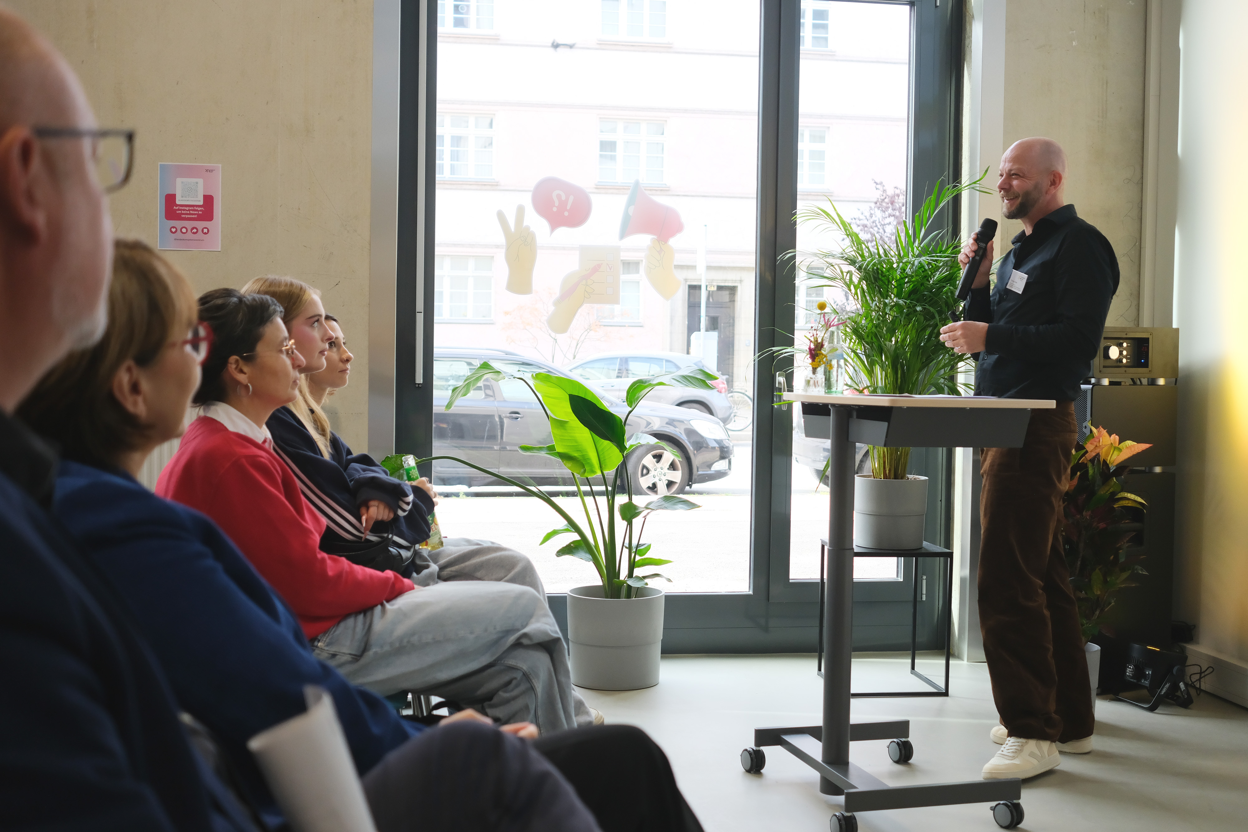 Ein Mann steht an einem Rednerpult und spricht in ein Mikrofon. Er trägt einen blauen Anzug und gestikuliert. Im Hintergrund sind große Fenster mit Pflanzen und Autos sichtbar.