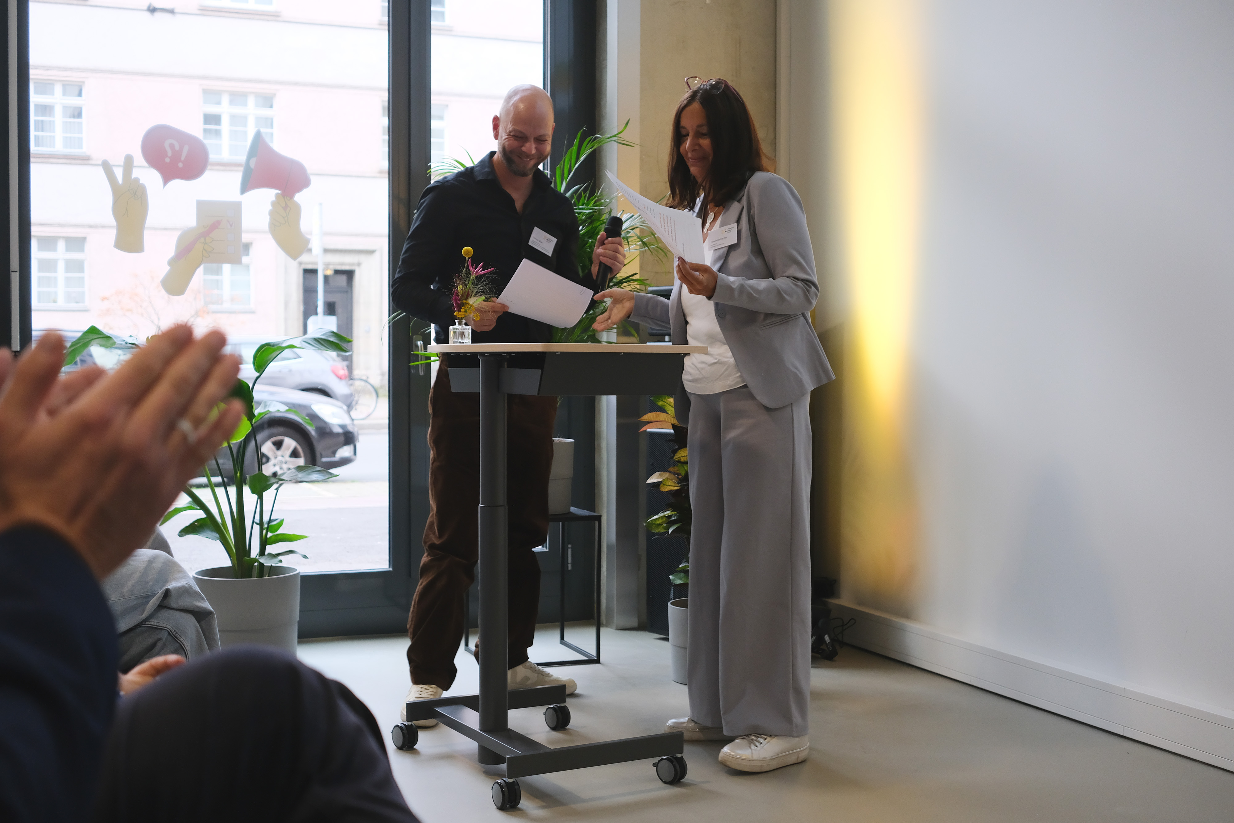Ein Mann und eine Frau stehen an einem Rednerpult und schauen sich Notizen an. Der Mann hält ein Mikrofon, während die Frau in einem grauen Anzug lächelt. Im Hintergrund sind Pflanzen und Fenster zu sehen.