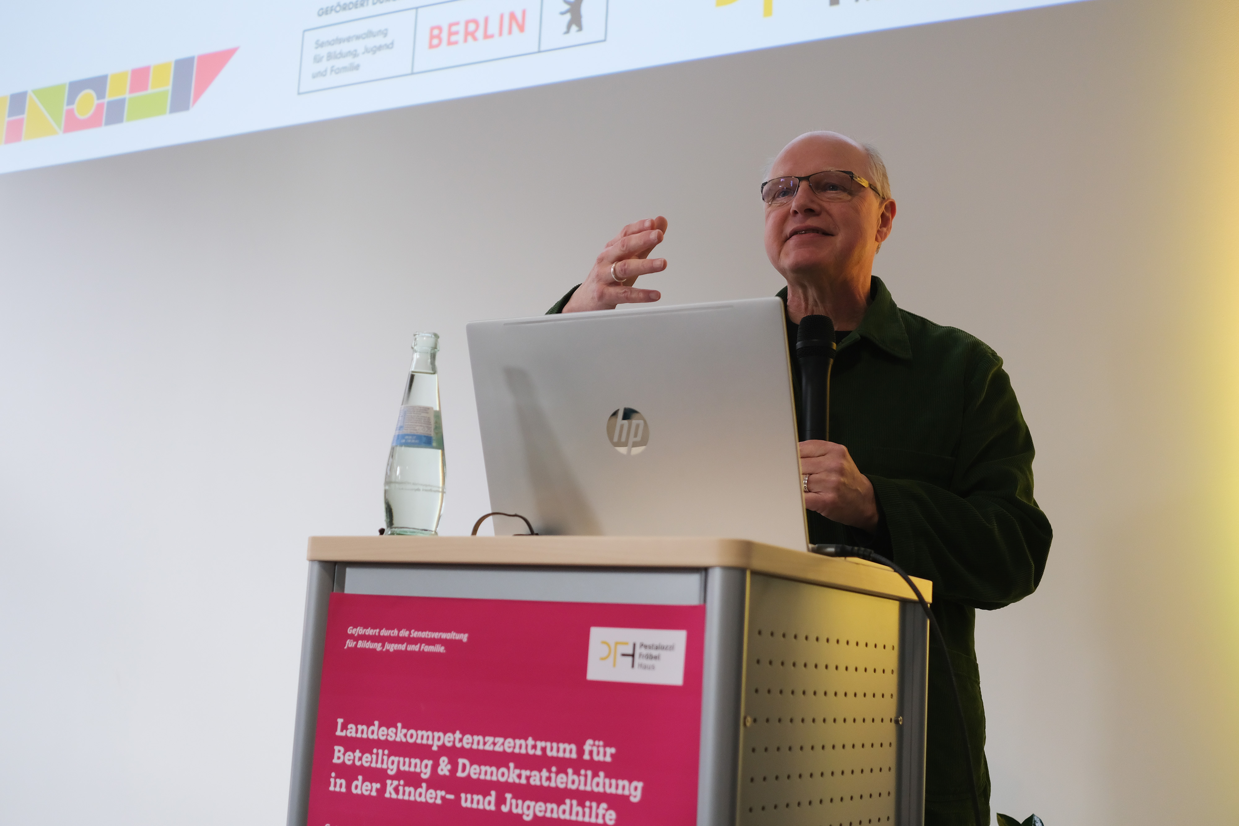 Ein Mann steht hinter einem Rednerpult mit einem Laptop und spricht. Er trägt eine grüne Jacke und hat eine Wasserflasche neben sich. Im Hintergrund ist eine Präsentation sichtbar, die Informationen über Demokratiebildung zeigt.