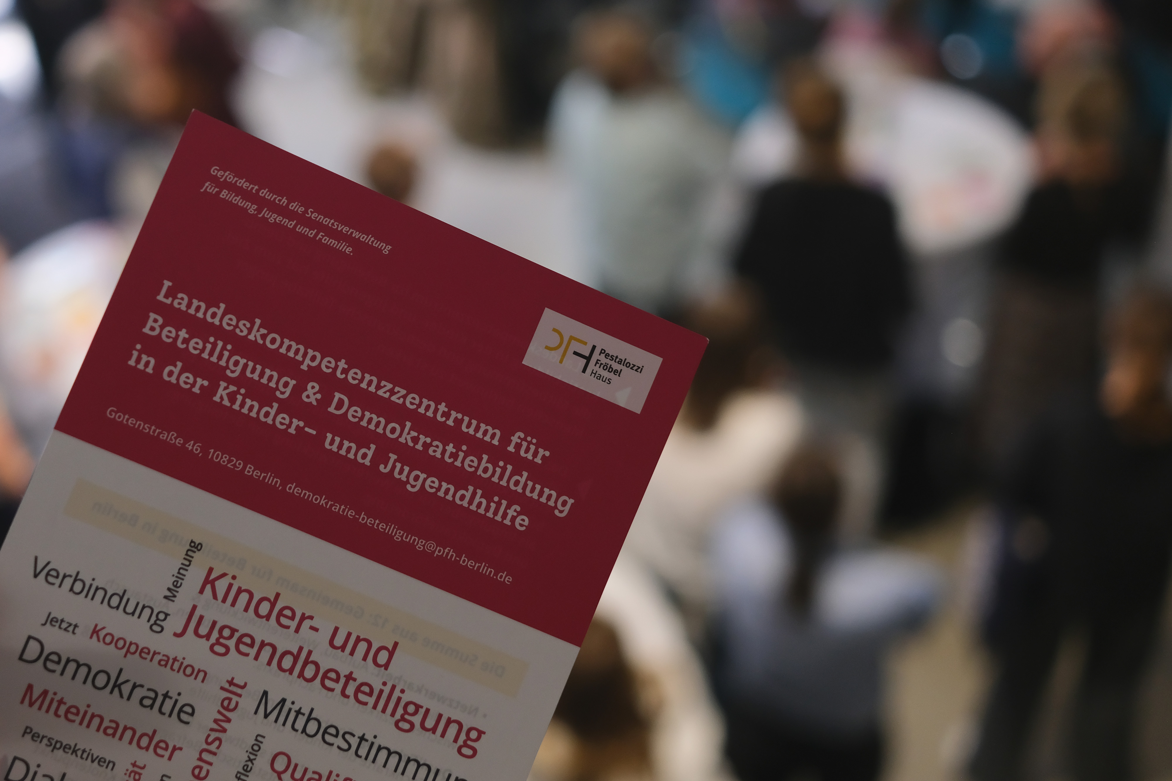 Eine Hand hält ein Informationsblatt mit dem Titel „Landeskompetenzzentrum für Beteiligung & Demokratiebildung in der Kinder- und Jugendhilfe“. Im Hintergrund sind unscharfe Personen zu sehen, die an einem Event teilnehmen.