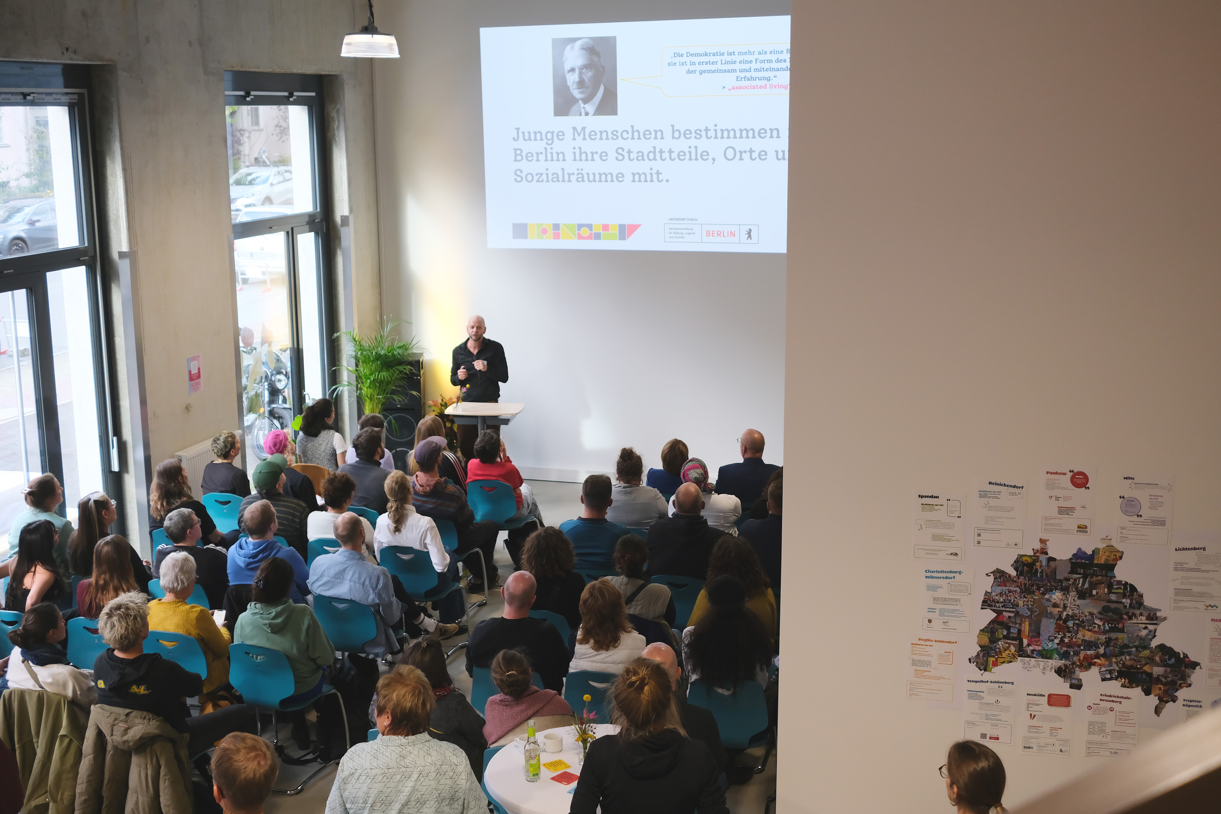 Eine Gruppe von Menschen sitzt in einem Raum und hört einem Vortrag zu. Ein Mann steht am Rednerpult und spricht, während im Hintergrund eine Präsentation an einer Wand gezeigt wird.