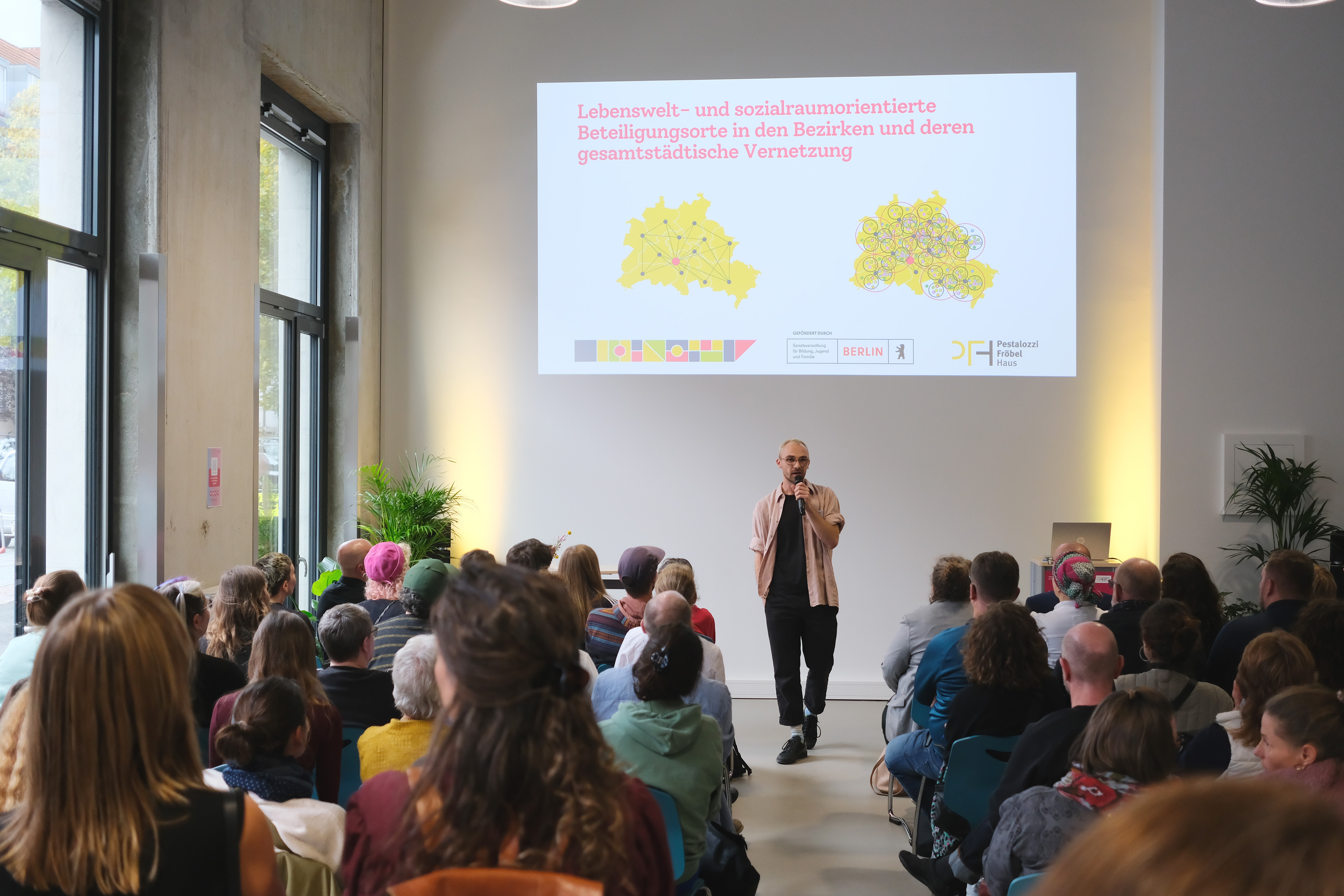 Ein Mann steht auf einer Bühne und spricht zu einem Publikum. Im Hintergrund sind zwei Karten mit Netzwerken in Berlin zu sehen. Die Zuschauer sitzen auf Stühlen und schauen aufmerksam zu.