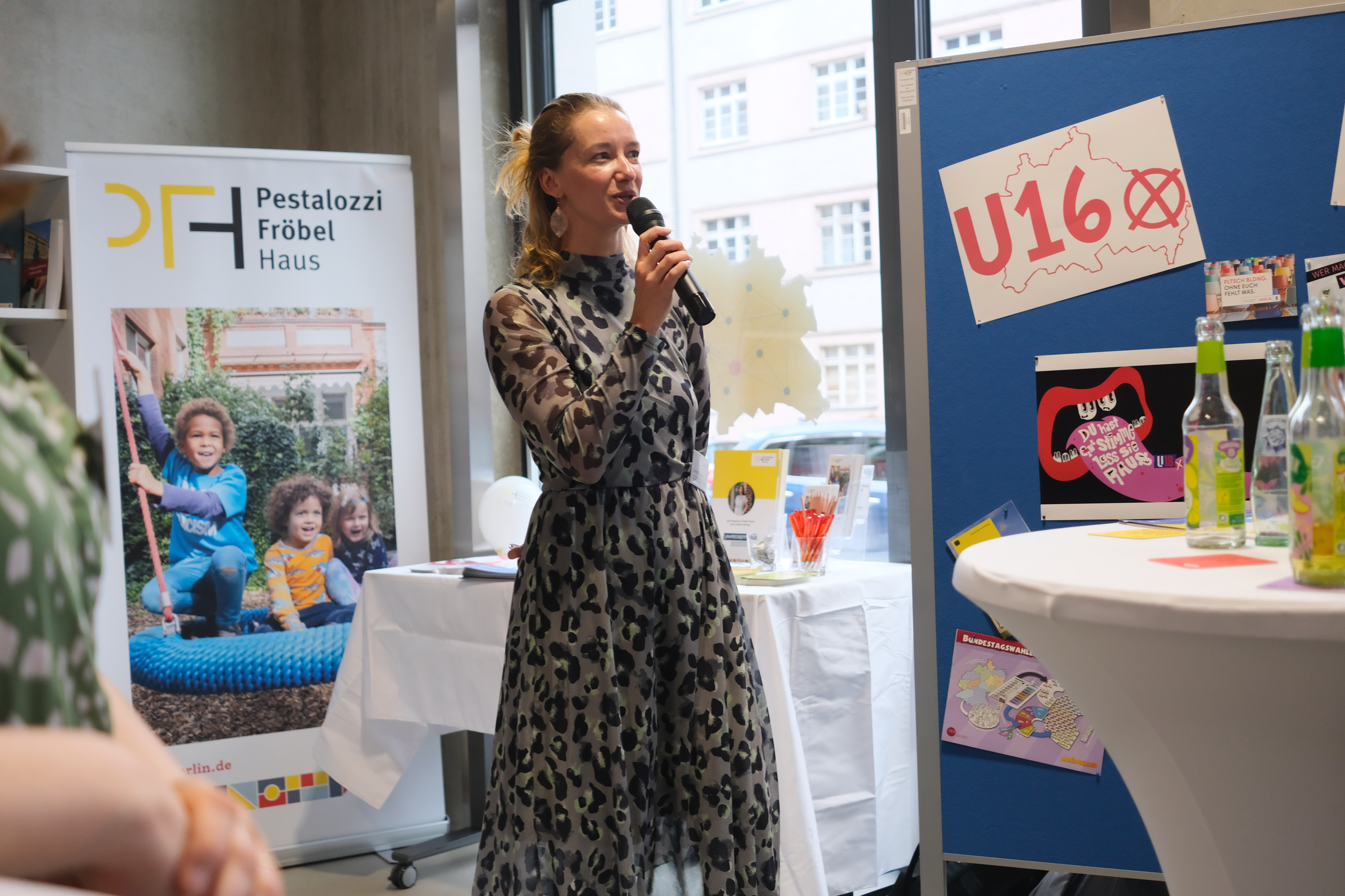Eine Frau in einem gemusterten Kleid hält ein Mikrofon und spricht. Im Hintergrund sind ein Banner mit dem Logo „Pestalozzi Fröbel Haus“ und ein Tisch mit Informationsmaterialien sichtbar.
