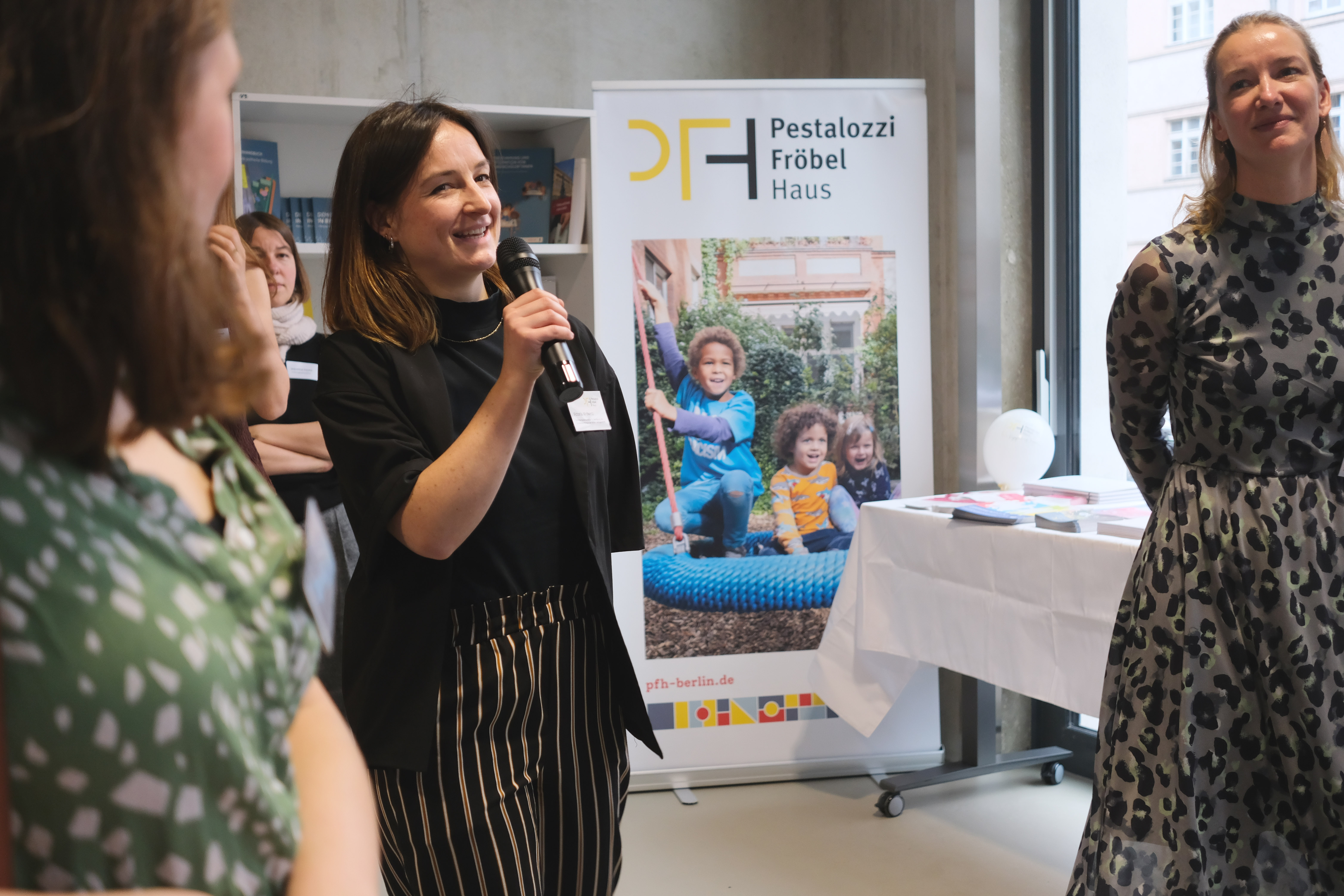 Eine Gruppe von Menschen applaudiert in einem Raum. Einige Personen stehen, während andere sitzen. Im Hintergrund sind Pflanzen und ein Banner mit dem Logo „Pestalozzi Fröbel Haus“ zu sehen.