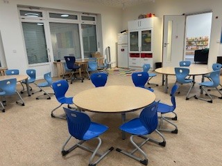 : Ein Raum mit einem großen runden Tisch und blauen Stühlen, der hell erleuchtet ist. Im Hintergrund sind Fenster und Regale mit verschiedenen Materialien sichtbar.