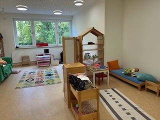   Bild 8: Ein Raum mit Regalen voller Spielzeug und Materialien. In der Mitte steht ein kleiner Tisch mit einem Stuhl, während im Hintergrund ein Bereich mit einem Spielhaus sichtbar ist.