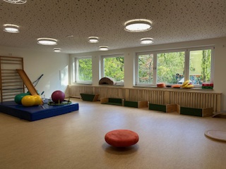 Ein heller Raum mit großen Fenstern, der mit verschiedenen Sportgeräten ausgestattet ist. Auf dem Boden liegen ein runder Sitzpuff und mehrere Bälle.