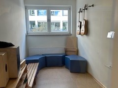  Ein Raum mit einer Holzbank und zwei Gitarren an der Wand. Der Raum ist hell und freundlich gestaltet, mit einem Fenster, das Tageslicht hereinlässt.