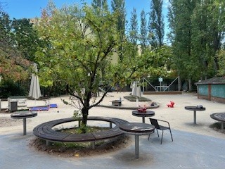 Ein Außenbereich mit einem großen Baum in der Mitte, umgeben von Spielgeräten, Tischen und Sitzgelegenheiten. 