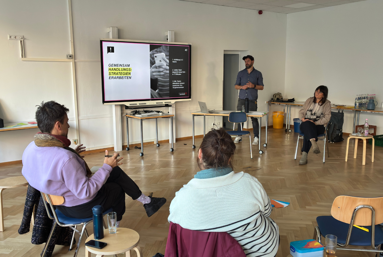Ein Mann und eine Frau stehen als Referent*innen neben einer Präsentation. Eine kleine Gruppe Menschen sitzt vor ihnen und hört zu.  