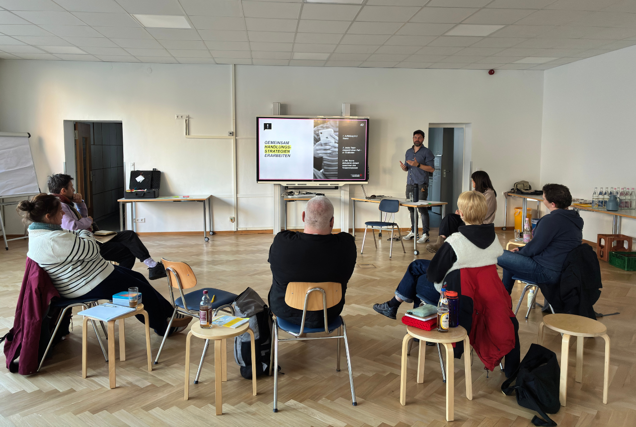 Ein Mann und eine Frau stehen als Referent*innen neben einer Präsentation. Eine Gruppe Menschen sitzt vor ihnen und hört zu.  