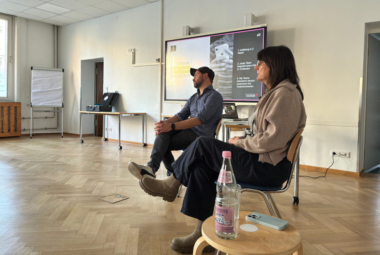 Ein Mann und eine Frau sitzen als Referent*innen neben einer Präsentation und hören aufmerksam zu. 