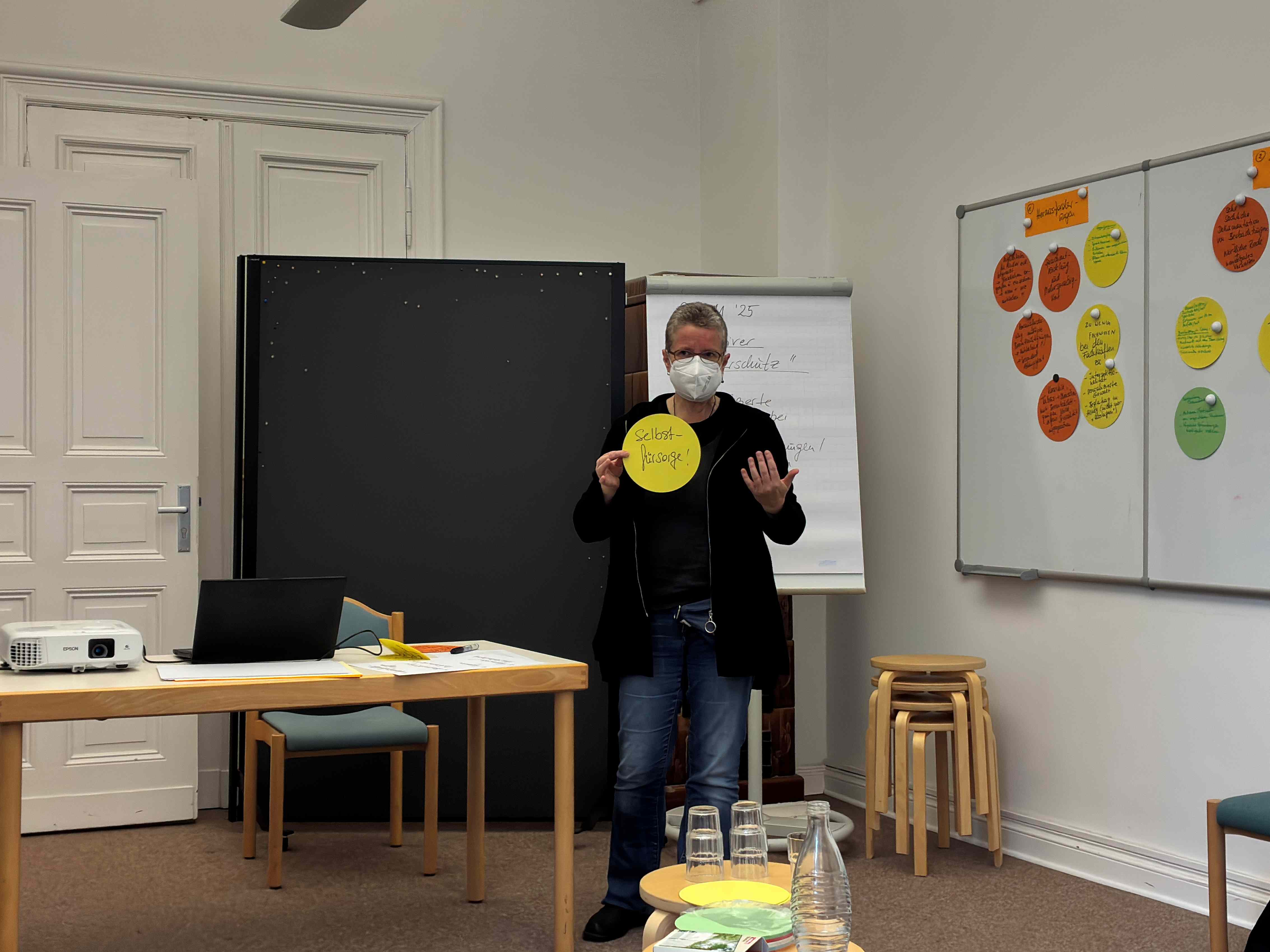 Die Person steht erneut und hält einen gelben Zettel in der Hand auf dem "Selbstfürsorge!" steht. Im Hintergrund sind die bunten Zettel an der Wand.