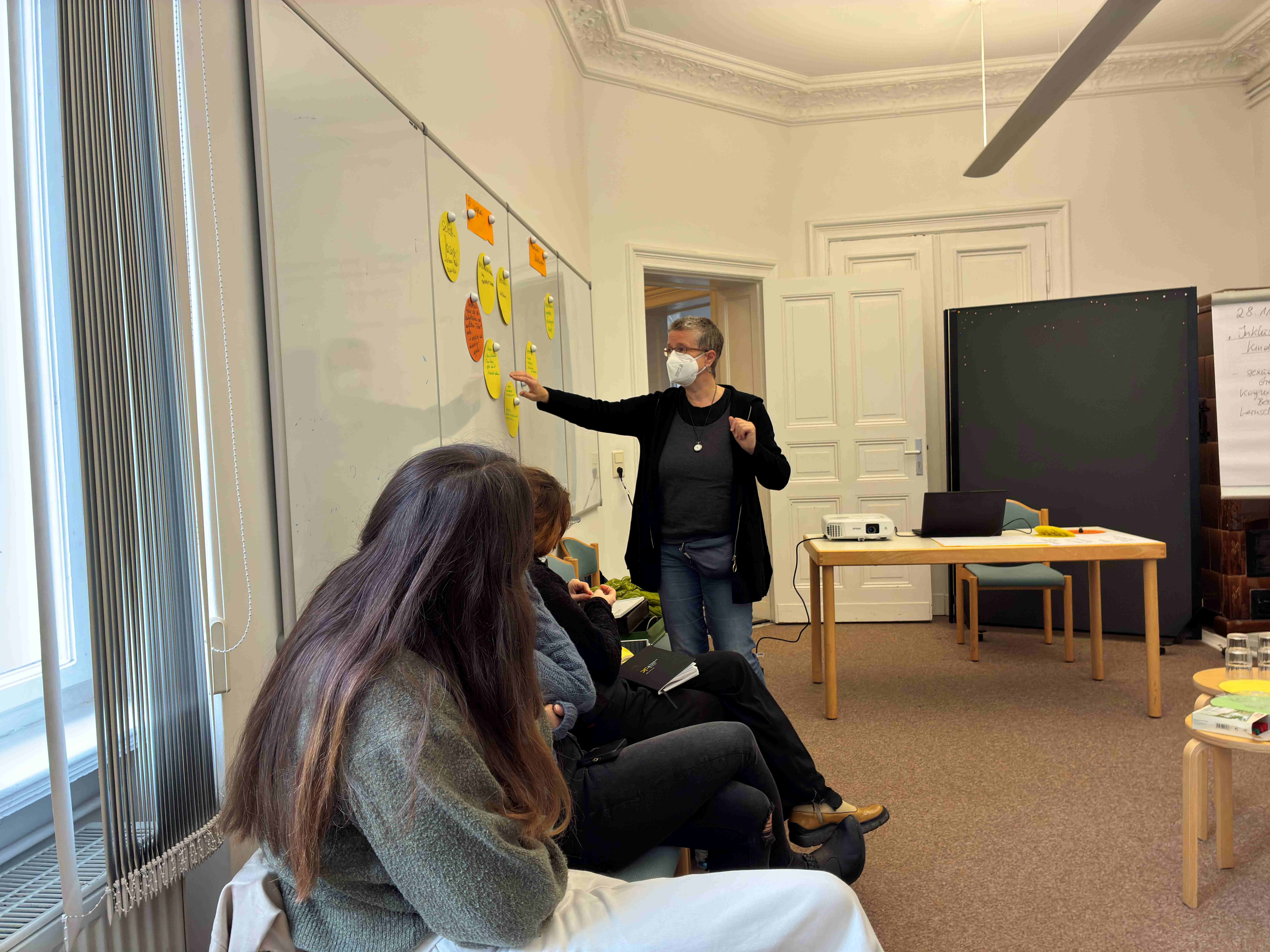Die selbe Person wie im vorherigen Bild zeigt auf die Wand mit bunten Zetteln. Die sitzenden Personen schauen in ihre Richtung. Der Raum ist hell und freundlich.