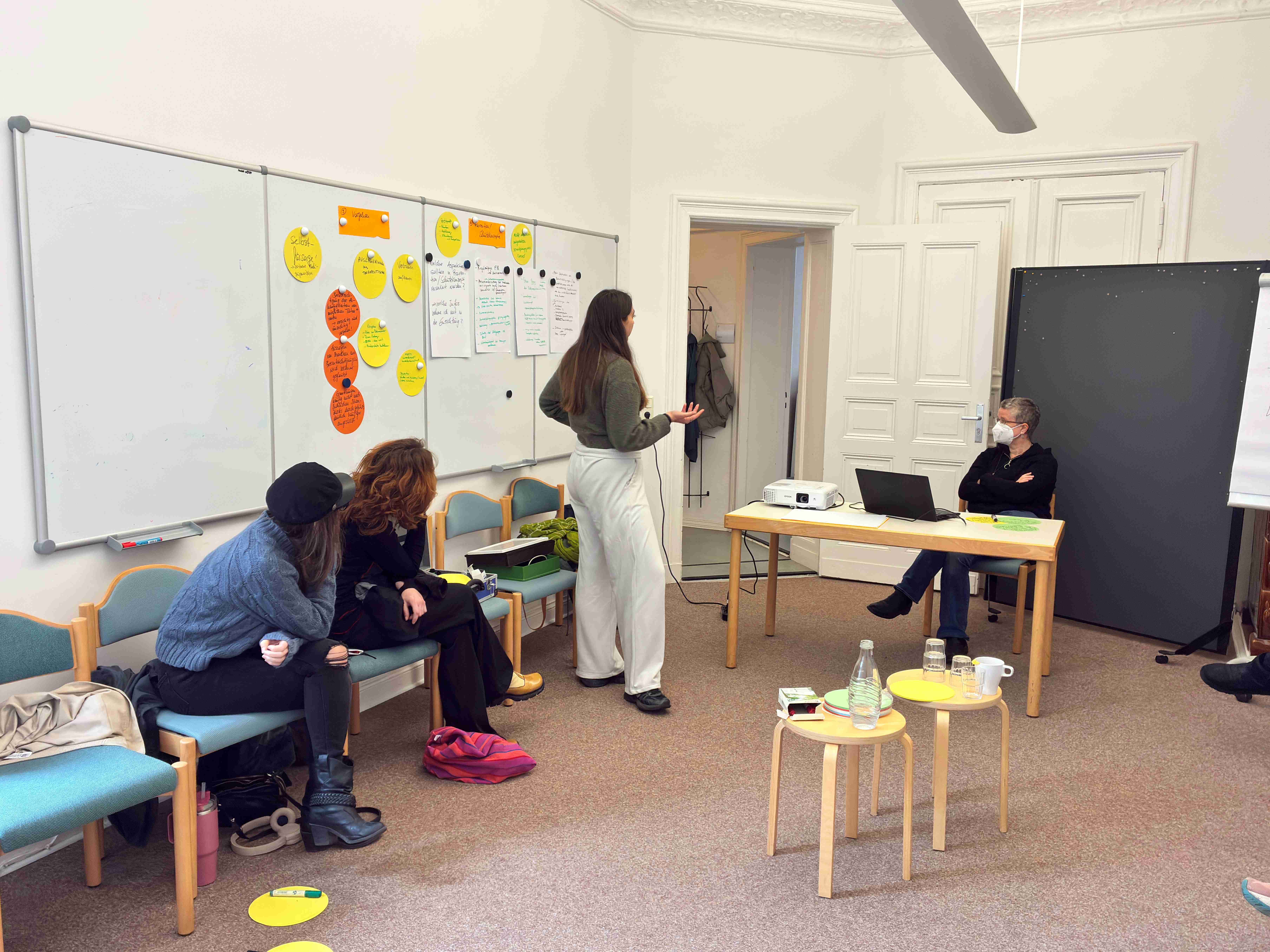 Eine Gruppe von vier Personen in einem Raum. Eine Person steht und spricht, während die anderen sitzen und zuhören. An der Wand sind mehrere bunte Zettel und Notizen angebracht. Auf einem Tisch steht ein Laptop und ein Projektor.