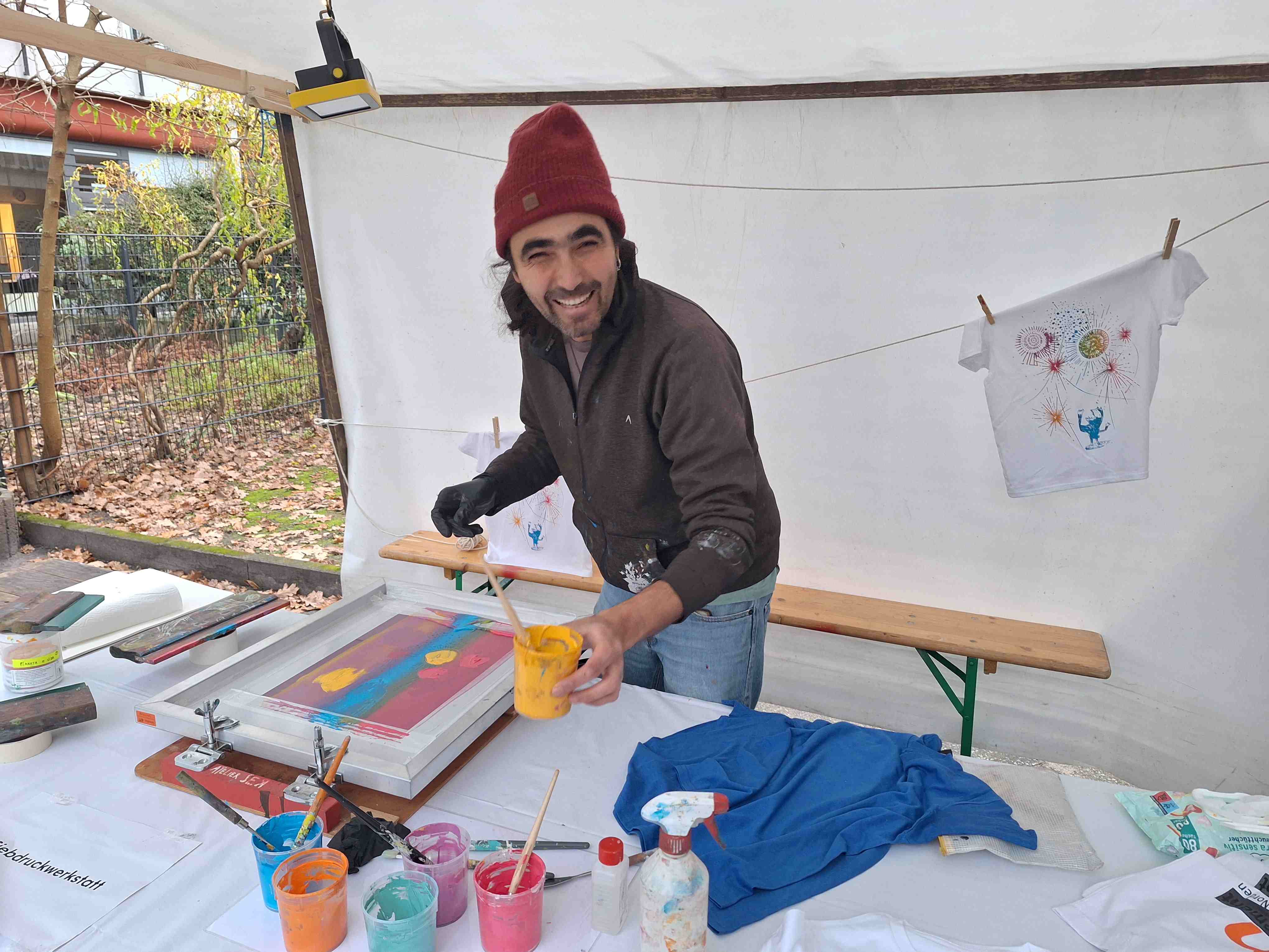 Ein Mann steht hinter einem Stand und lächelt in die Kamera. Er hält einen Pinsel in der Hand und bemalt ein T-Shirt mit bunten Farben.