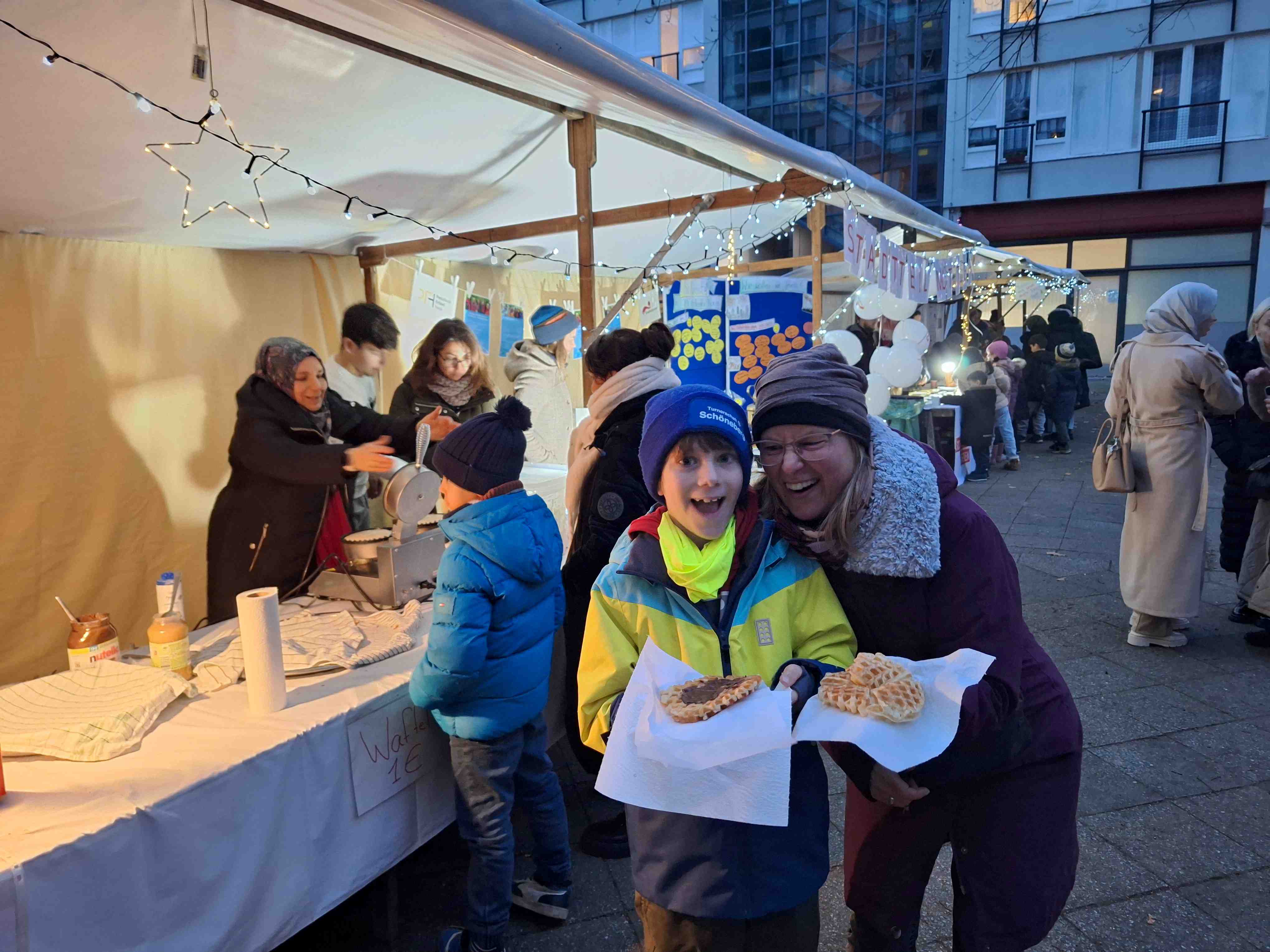 Ein Kind und eine Frau halten Waffeln in der Hand und lächeln in die Kamera. Im Hintergrund sieht man winterlich geschmückte Marktstände.