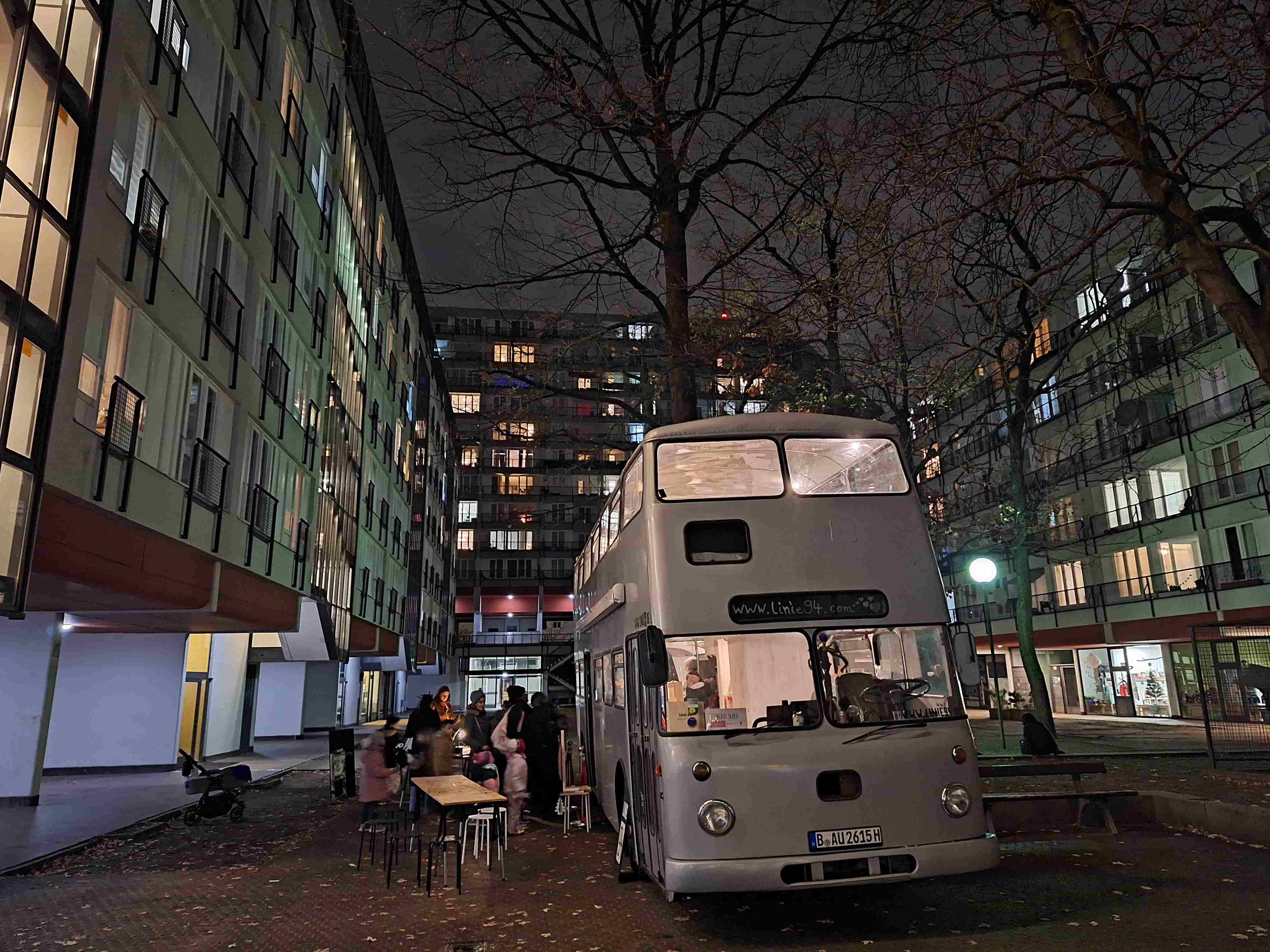 Ein zweistückiger silberner Bus der Linie94 steht am Straßenrand. Es ist dunkel und die Lichter des Buses leuchten. Eine kleine Gruppe Personen sitzt an Tischen vor dem Bus.