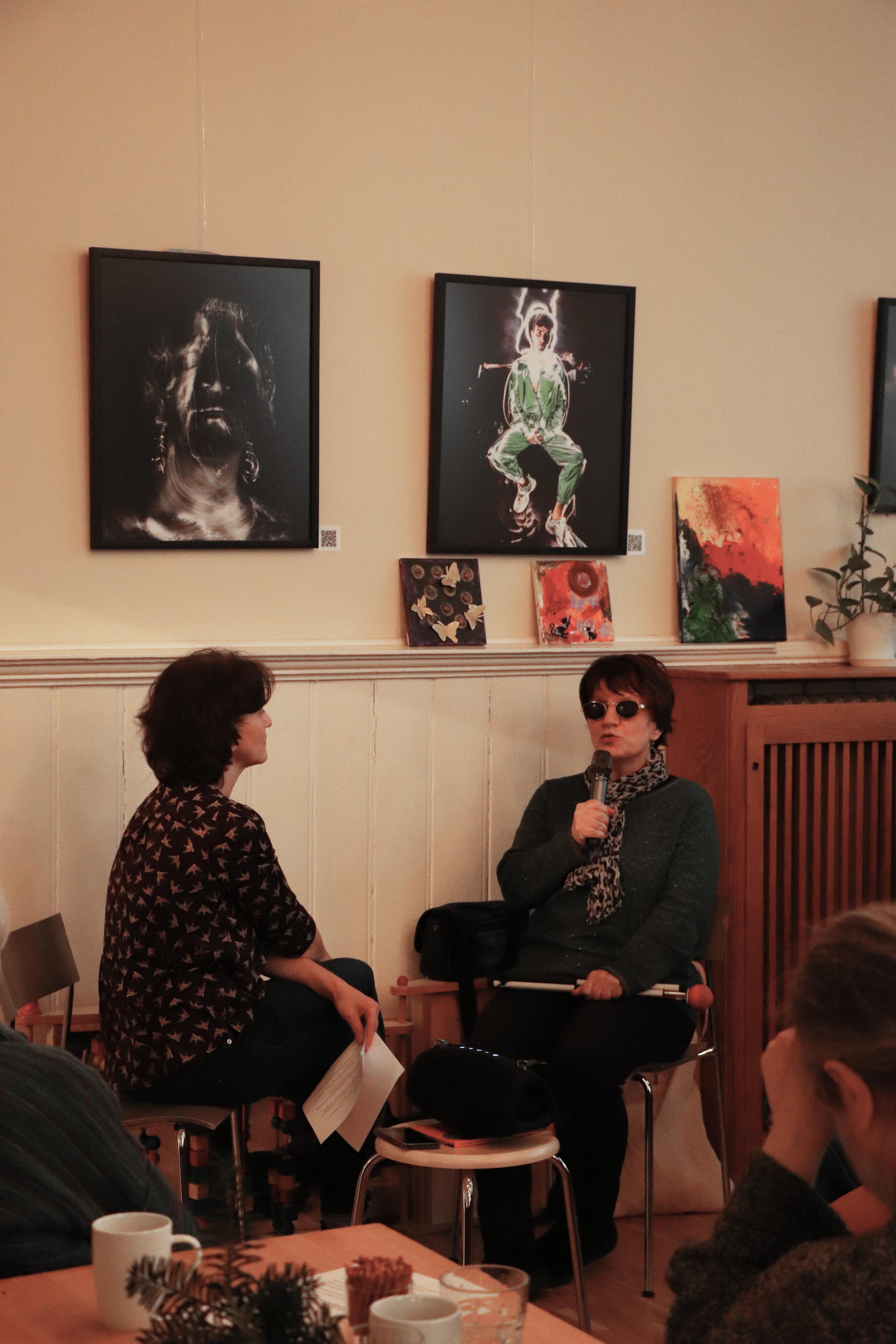 Zwei Frauen sitzen sich gegenüber. Die eine Frau hält einen Zettel in der Hand, die andere Frau hält ein Mikrophon und spricht. Sie trägt eine Sonnenbrille. Im Hintergrund hängen zwei Kunstwerke an der Wand.