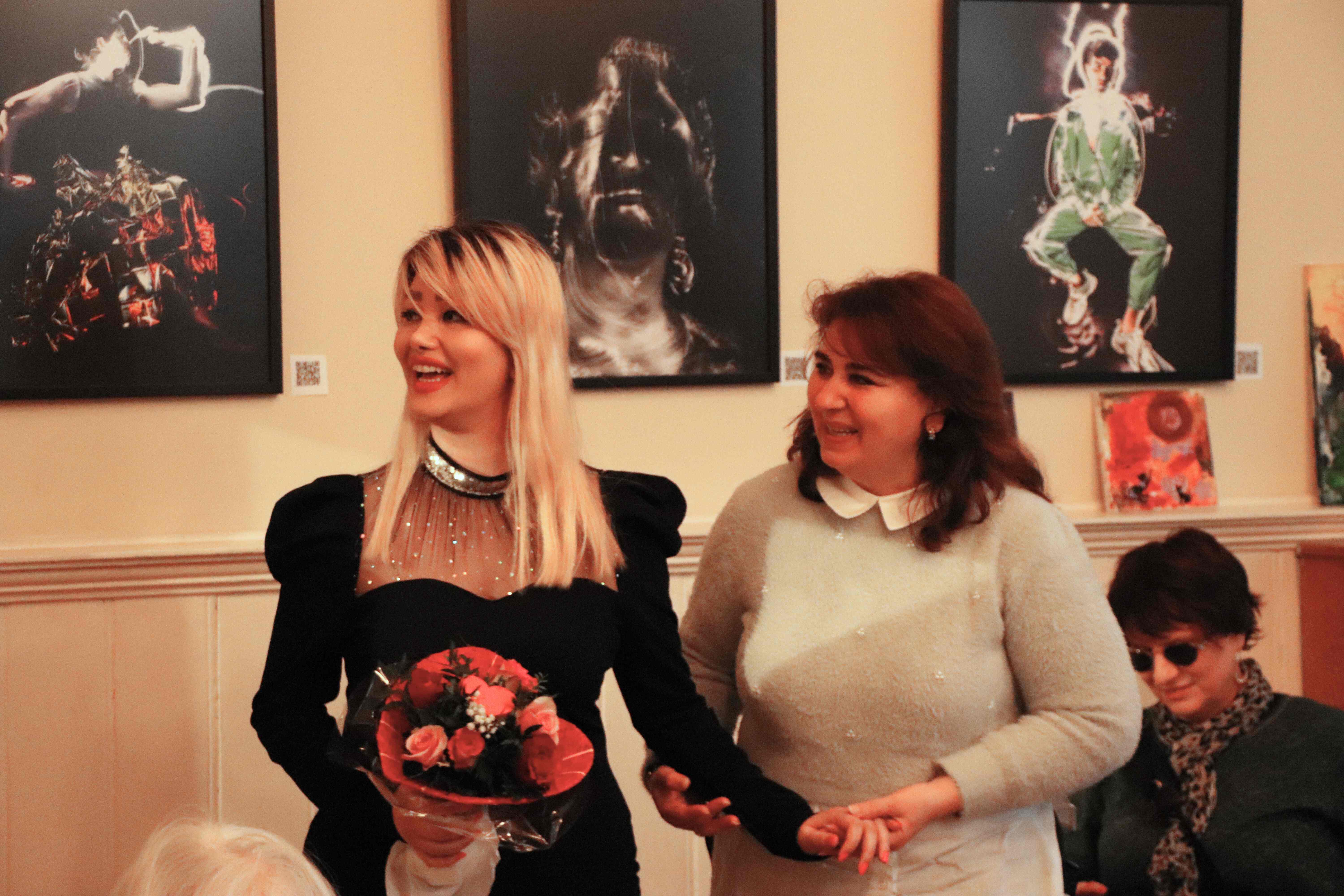 Zwei Frauen stehen nebeneinander und lächeln. Die eine Frau hält einen Blumenstrauß in der Hand. Im Hintergrund hängen drei Kunstwerke an der Wand.