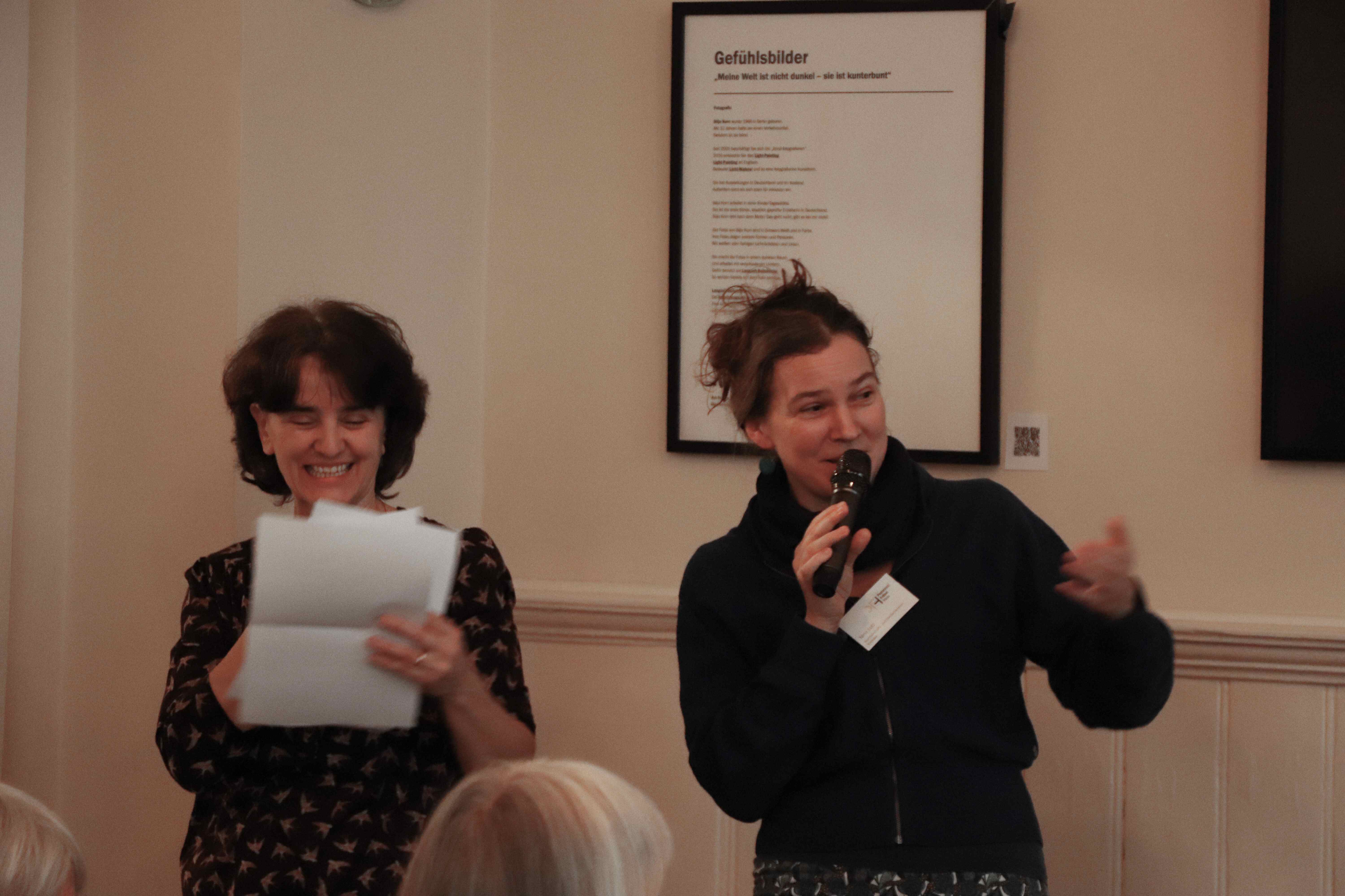 Zwei Frauen stehen nebeneinander. Eine hält einen Zettel in der Hand und lächelt, während die andere durch ein Mikrofon zu einem Publikum spricht.