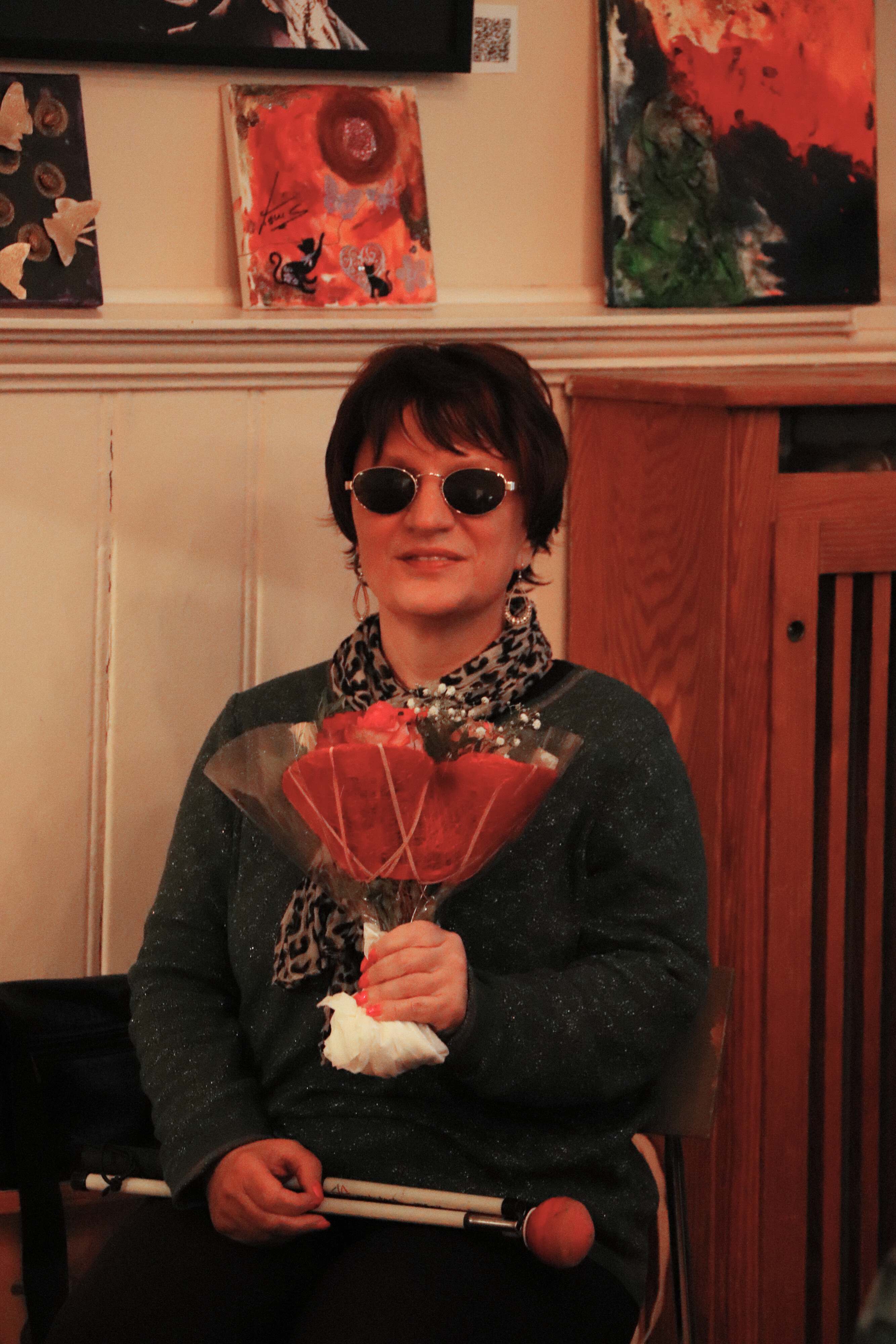 Eine Frau sitzt auf einem Stuhl. Sie trägt eine Sonnenbrille, hält einen Blumenstrauß in der Hand und lächelt.