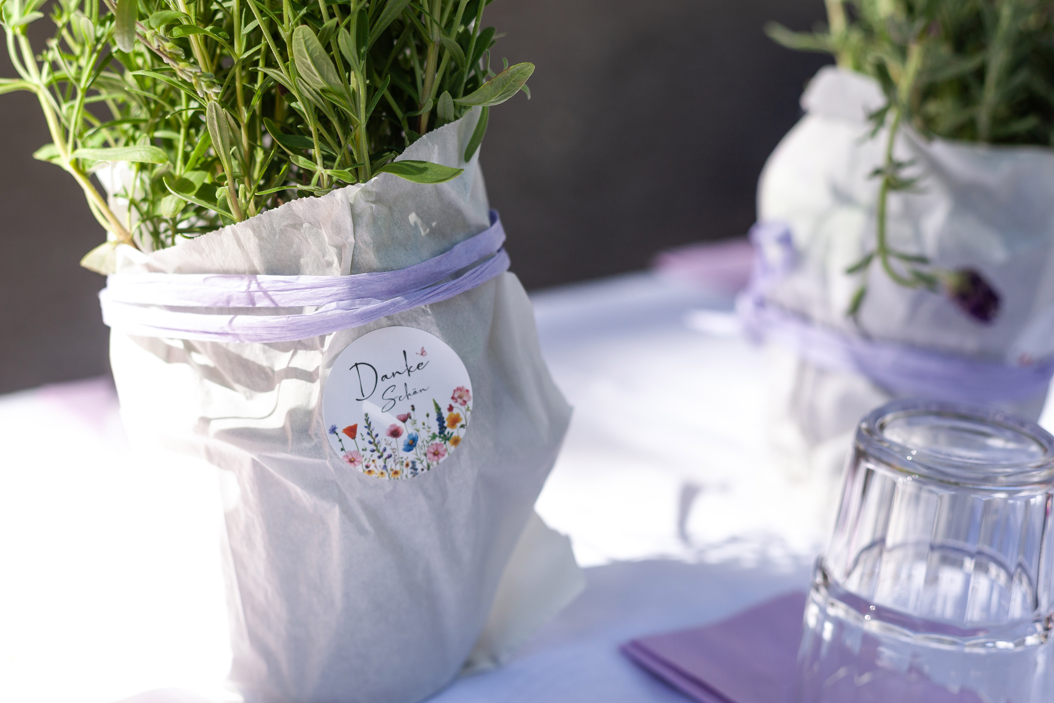 Tischdeko ist zu sehen. Kleine Lavendelpflanzen sind mit weißem Papier umhüllt und mit einer hell violetten Schleife umbunden. Verziert sind die mit einem Aufkleber mit der Aufschrift Danke.