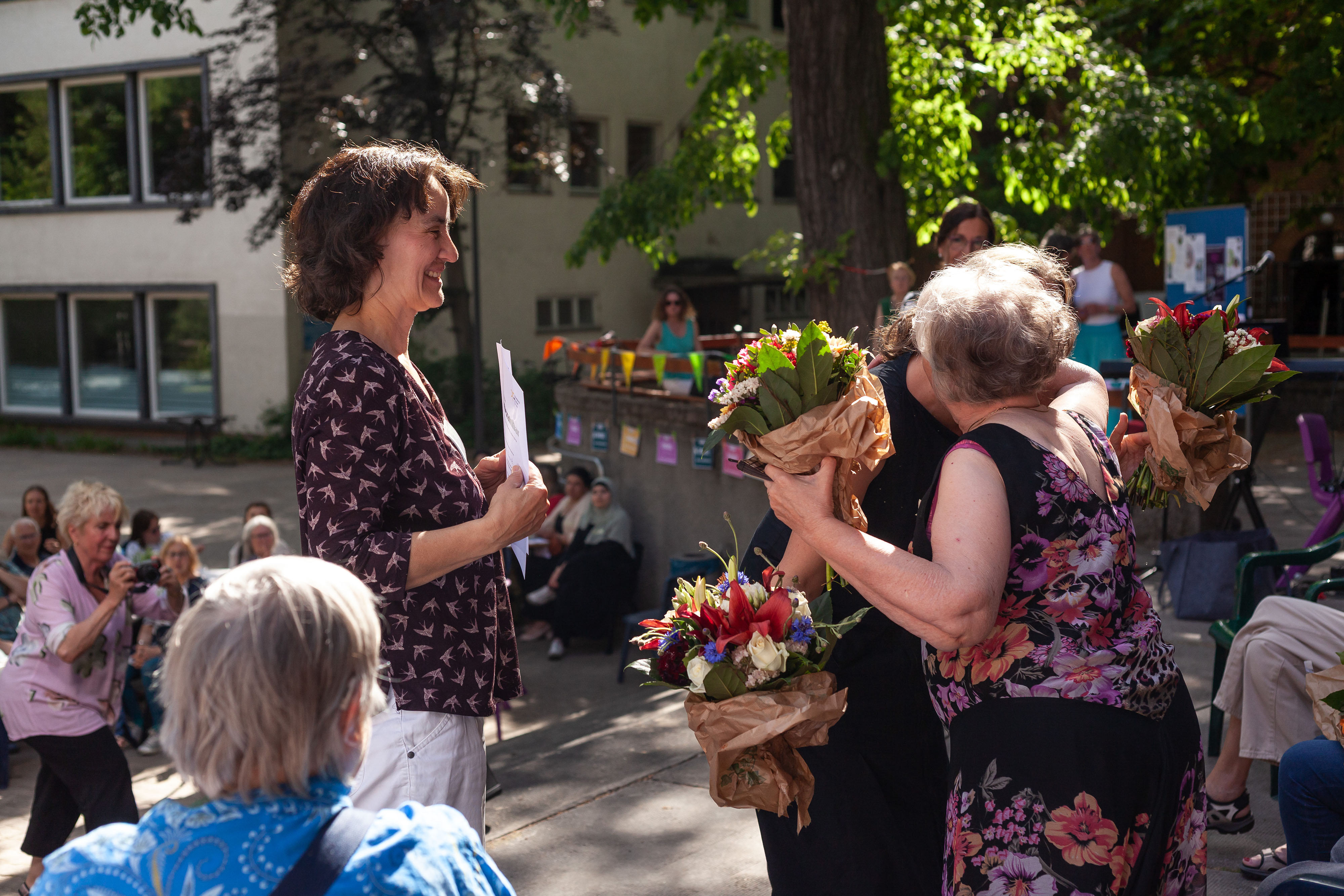 Gertraude Langbehn von Unbezahlbar und Nina Lutz aus der Kiezoase gratulieren einer Ehrenamtlichen mit einem Blumenstrauß und einer Urkunde.Nina Lutz umarmt die Ehrenamtliche.