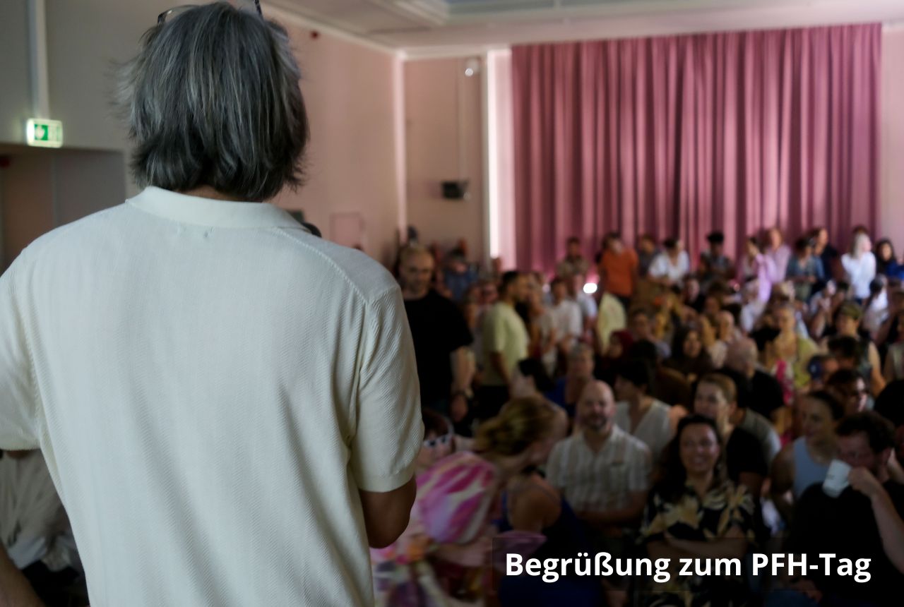 Blick von hinten auf eine Person, die vor einem vollen Saal spricht; viele Menschen sitzen und stehen, lächeln und schauen zur Bühne, rosafarbener Vorhang im Hintergrund.