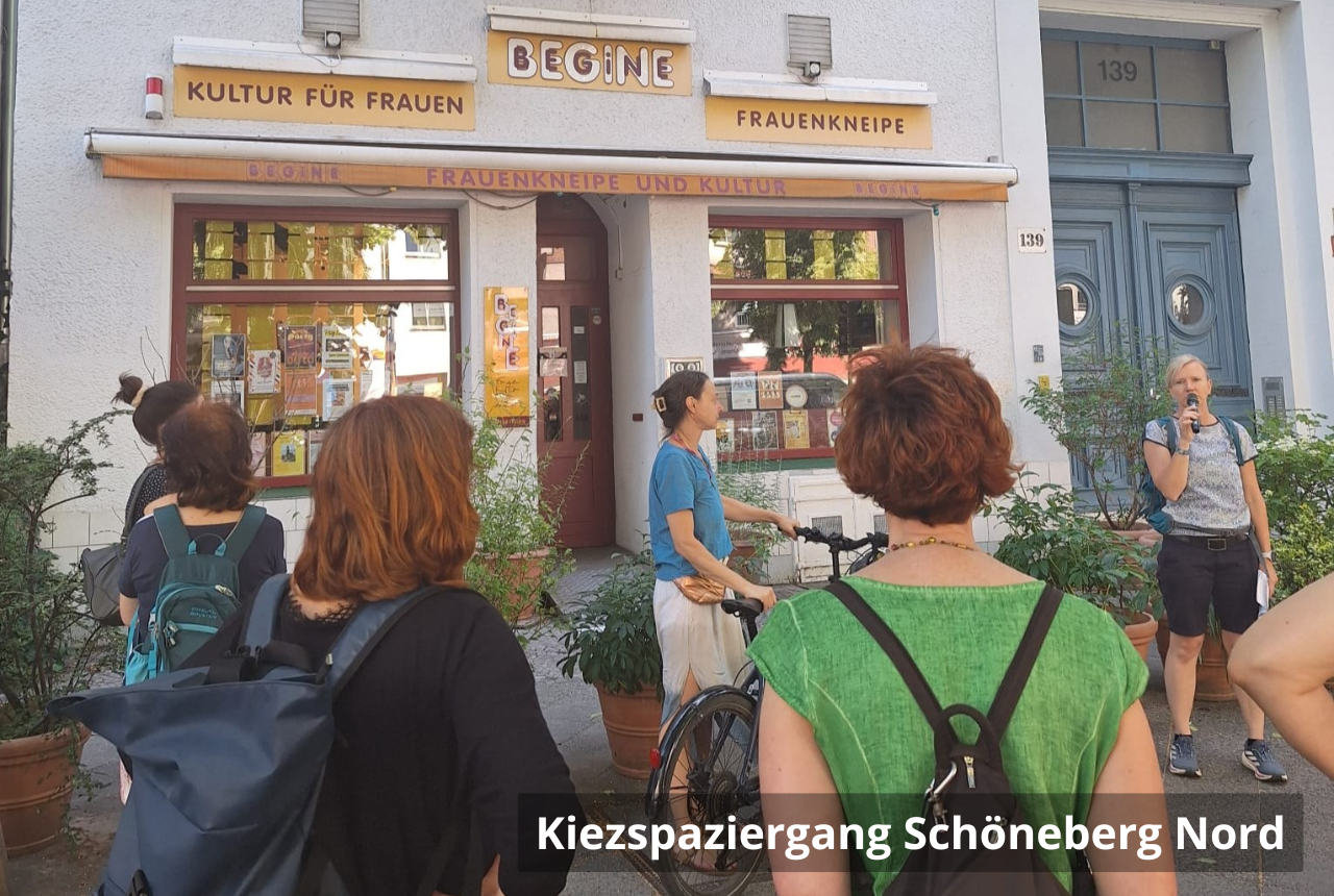Kiezspaziergang Schöneberg Nord: Sechs Personen stehen im Kreis auf dem Fußweg. Eine Frau spricht in ein Mikrofon, die anderen hören ihr zu. Im Hintergrund ist eine Kneipe mit einem Schild: Frauenkneipe Begine - Kultur für Frauen. 