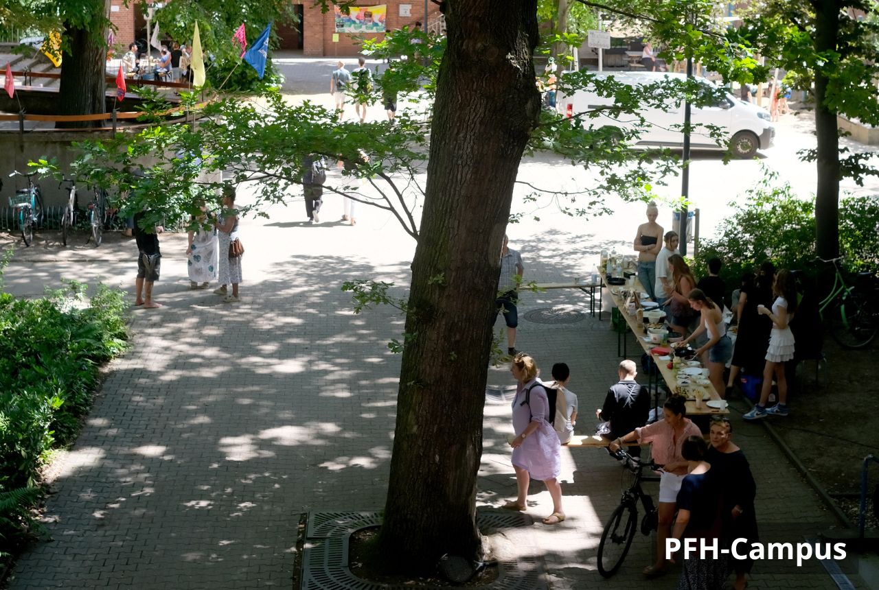 Schattenreicher Hof mit großem Baum in der Mitte. Rechts steht eine lange Biertisch-Garnitur, an der mehrere Menschen sprechen, sitzen und Essen/Getränke anrichten; eine Person kommt mit Fahrrad dazu. Links und im Hintergrund gehen weitere Personen über den gepflasterten Platz und bunte Fahnen hängen zwischen den Bäumen.