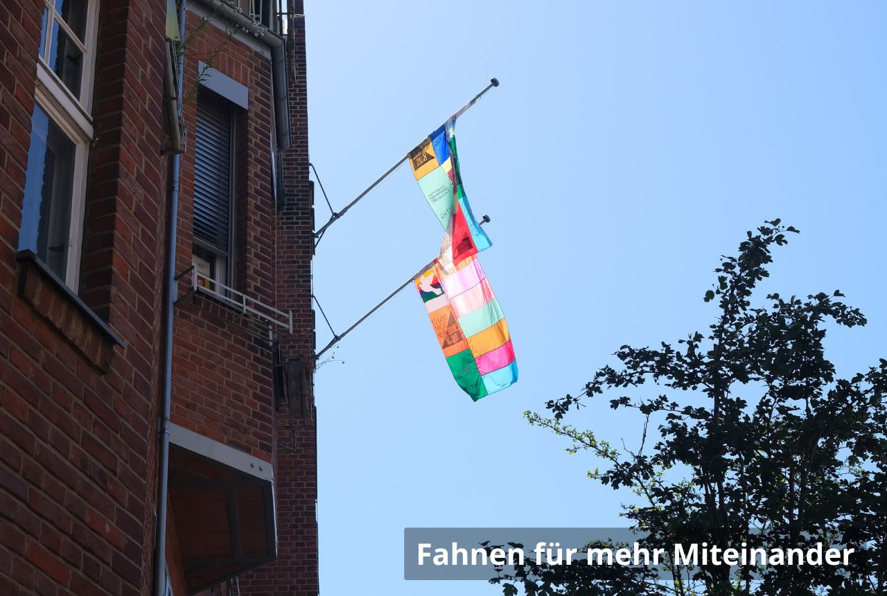 Bunte, aus Stoffstreifen zusammengesetzte Fahne weht an einer Hausfassade aus rotem Backstein. Die Flagge hängt an einem schrägen Mast über der Straße, vor klarem blauem Himmel; rechts ragen Zweige eines Baumes ins Bild.