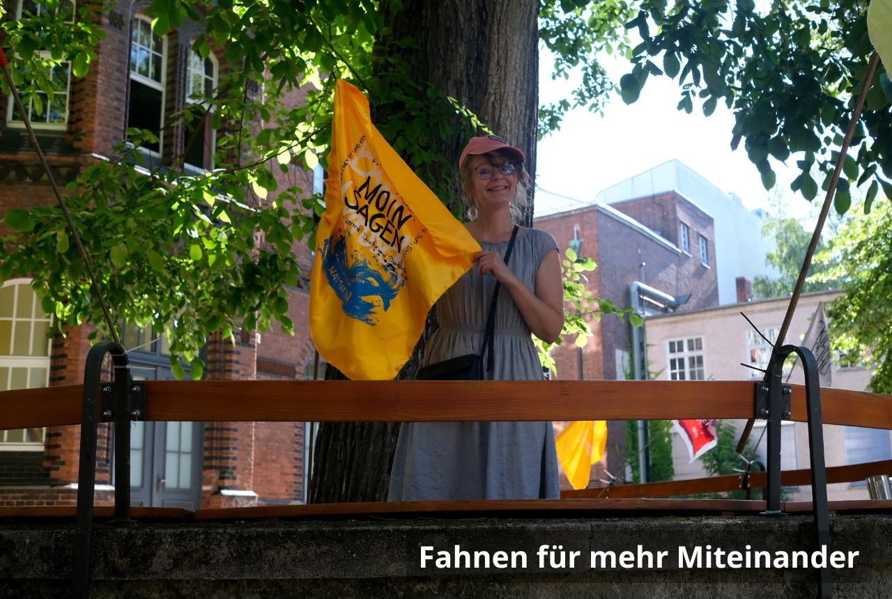 Eine Frau hält eine gelbe Flagge auf der "Moin" steht in die Höhe. Sie lächelt. Im Hintergrund ist ein Backsteingebäude, blauer Himmel und ein grüner Baum. 