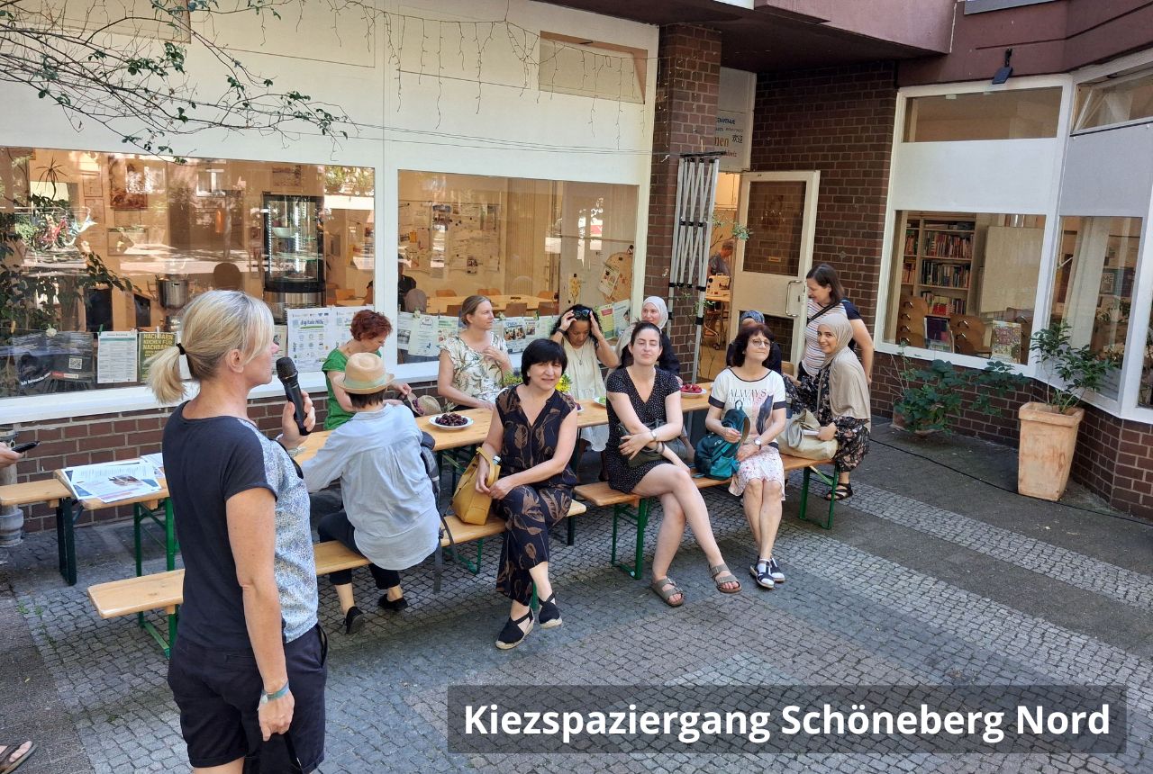  Gruppe von Menschen sitzt und steht auf Bänken vor einem Nachbarschaftstreff; eine Frau spricht mit Mikrofon, hinter ihr große Fenster mit Aushängen.