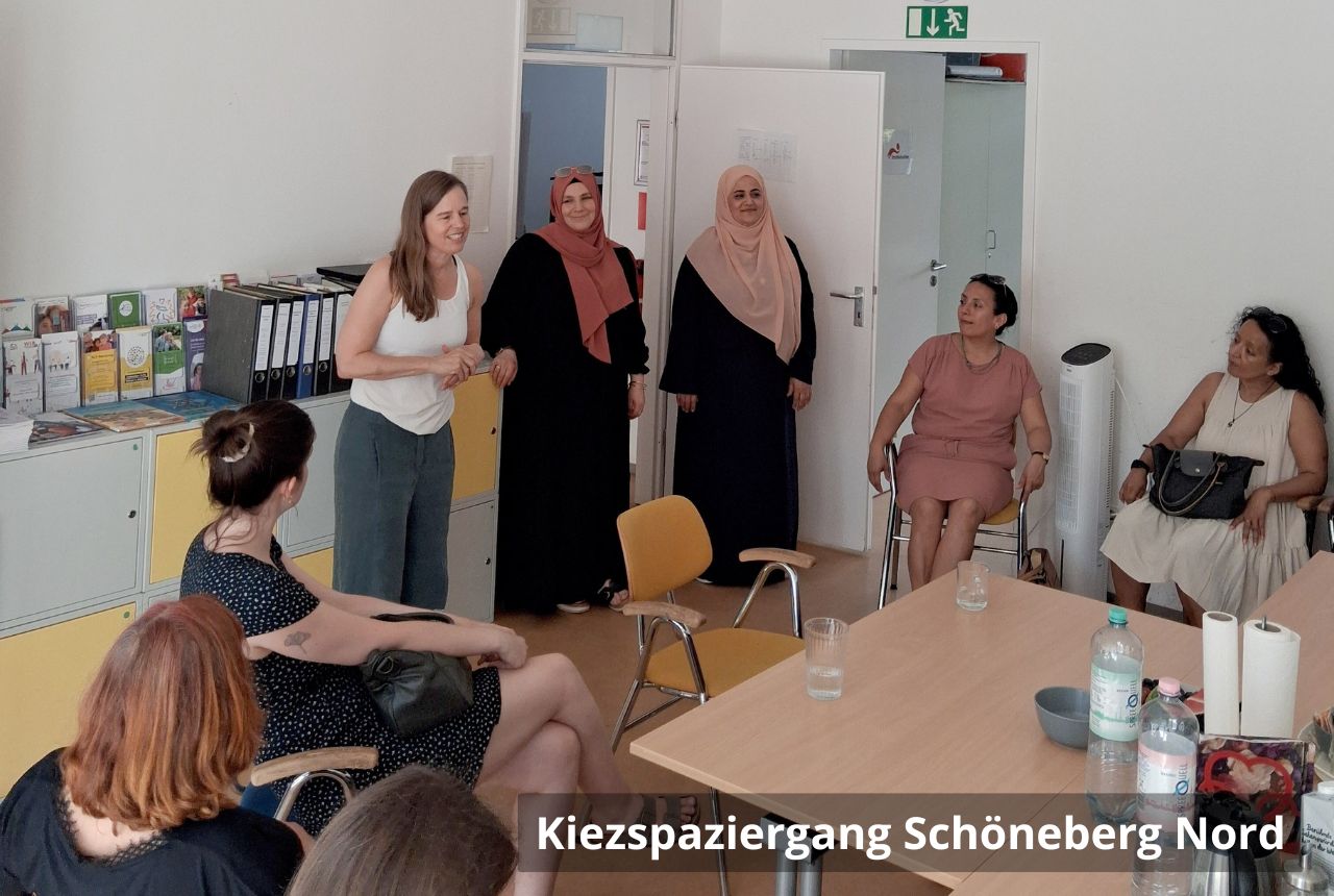  Mehrere Frauen sitzen und stehen in einem hellen Besprechungsraum; drei Personen vorn am Türbereich sprechen zu der Runde, auf dem Tisch stehen Wasserflaschen und Tassen.