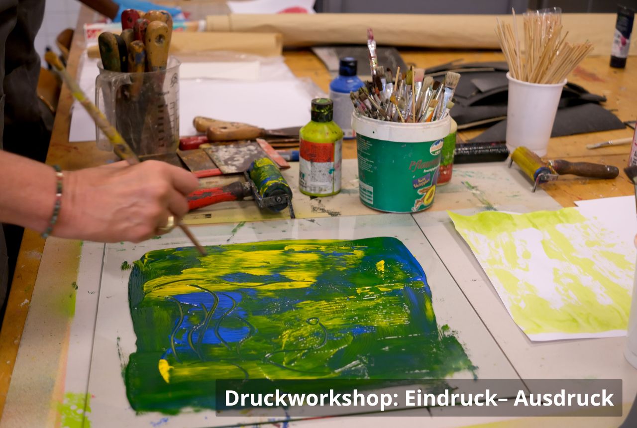 Person arbeitet an einem Werk­tisch voller Malutensilien. Auf einer Glasplatte werden mit Pinsel gelbe und blaue Farbe verteilt; daneben stehen Pinselbecher, Farben und verschiedene Werkzeuge.