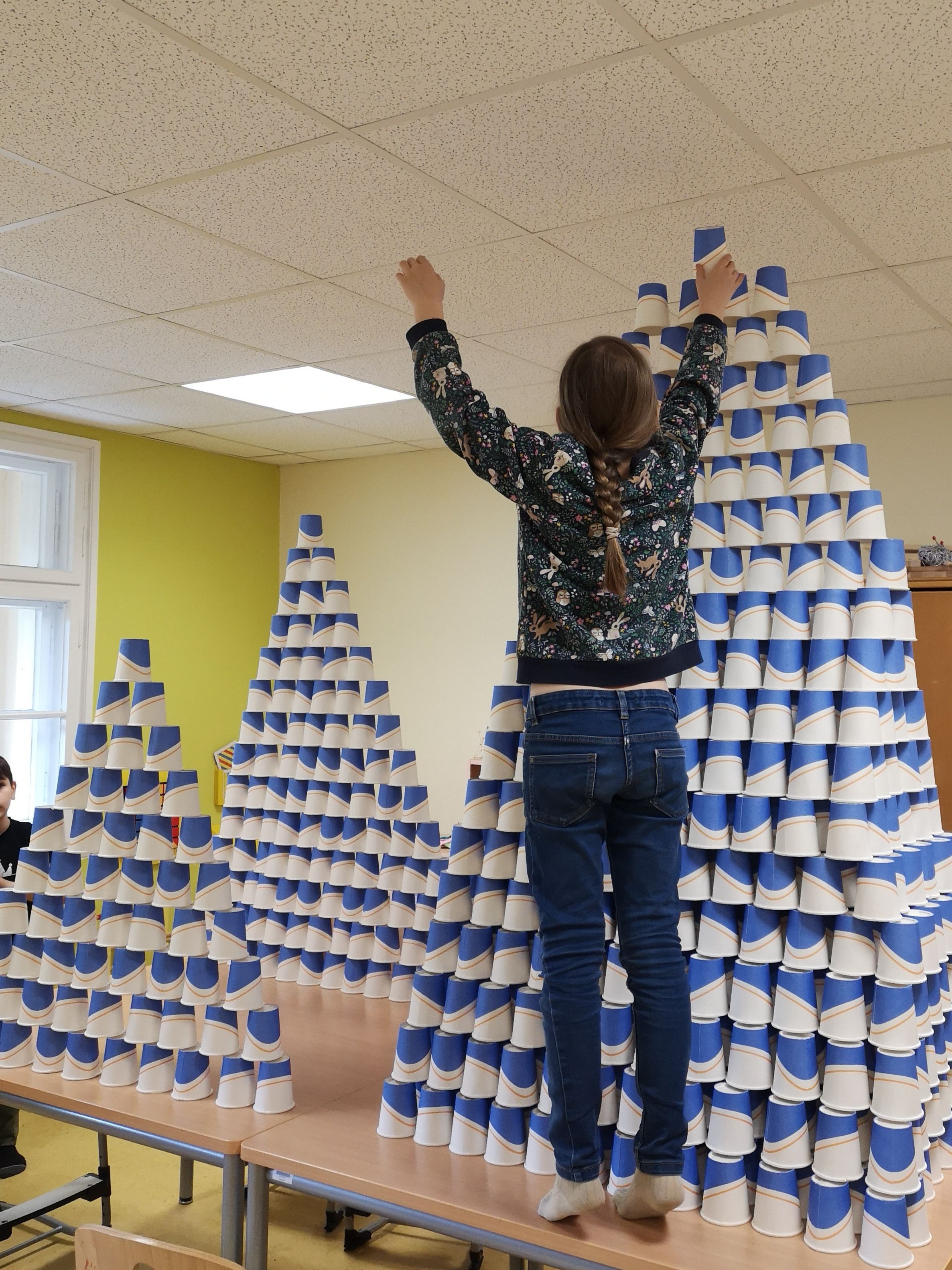 In einem Gruppenraum werden aus blau-weißen Pappbechern mehrere hohe Türme aufgebaut. Eine Person auf einem Tisch setzt einen Becher auf die Spitze einer großen Konstruktion.