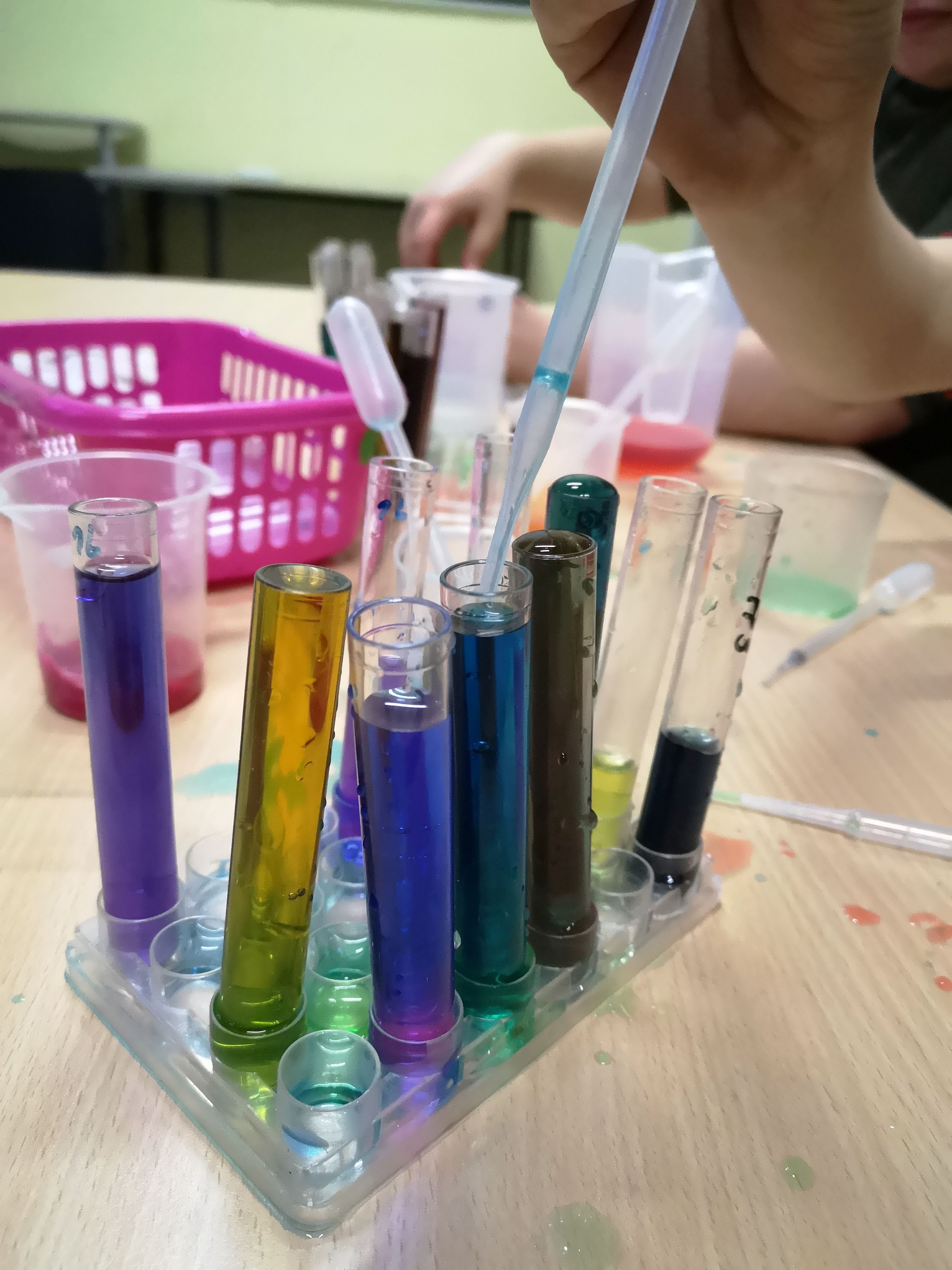 Bei einem Experiment werden farbige Flüssigkeiten mit einer Pipette in Reagenzgläser gefüllt. Die Reagenzgläser enthalten verschiedene Blau-, Grün-, Gelb- und Violetttöne und stehen in einem transparenten Halter auf einem Tisch.