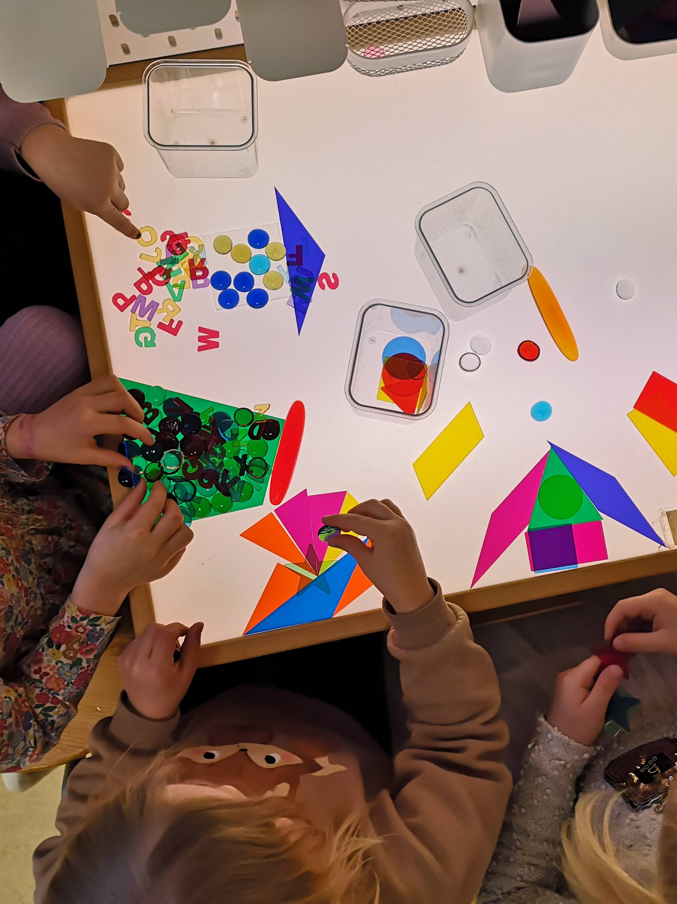 Kinder experimentieren an einem Leuchttisch mit bunten transparenten Formen, Buchstaben und Spielsteinen. Die Materialien werden sortiert und zu geometrischen Figuren und Mustern zusammengesetzt.